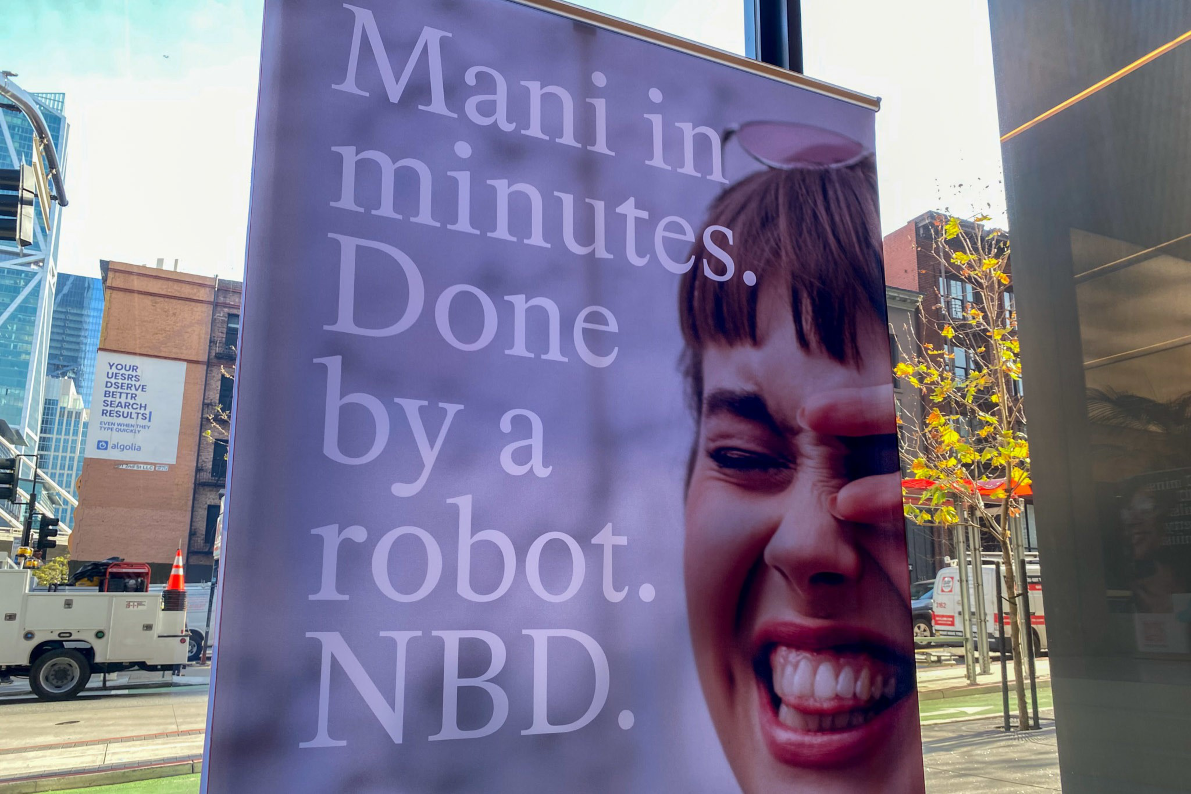 A large banner displays a close-up of a laughing woman with short hair and text reading "Mani in minutes. Done by a robot. NBD." in a city street setting.