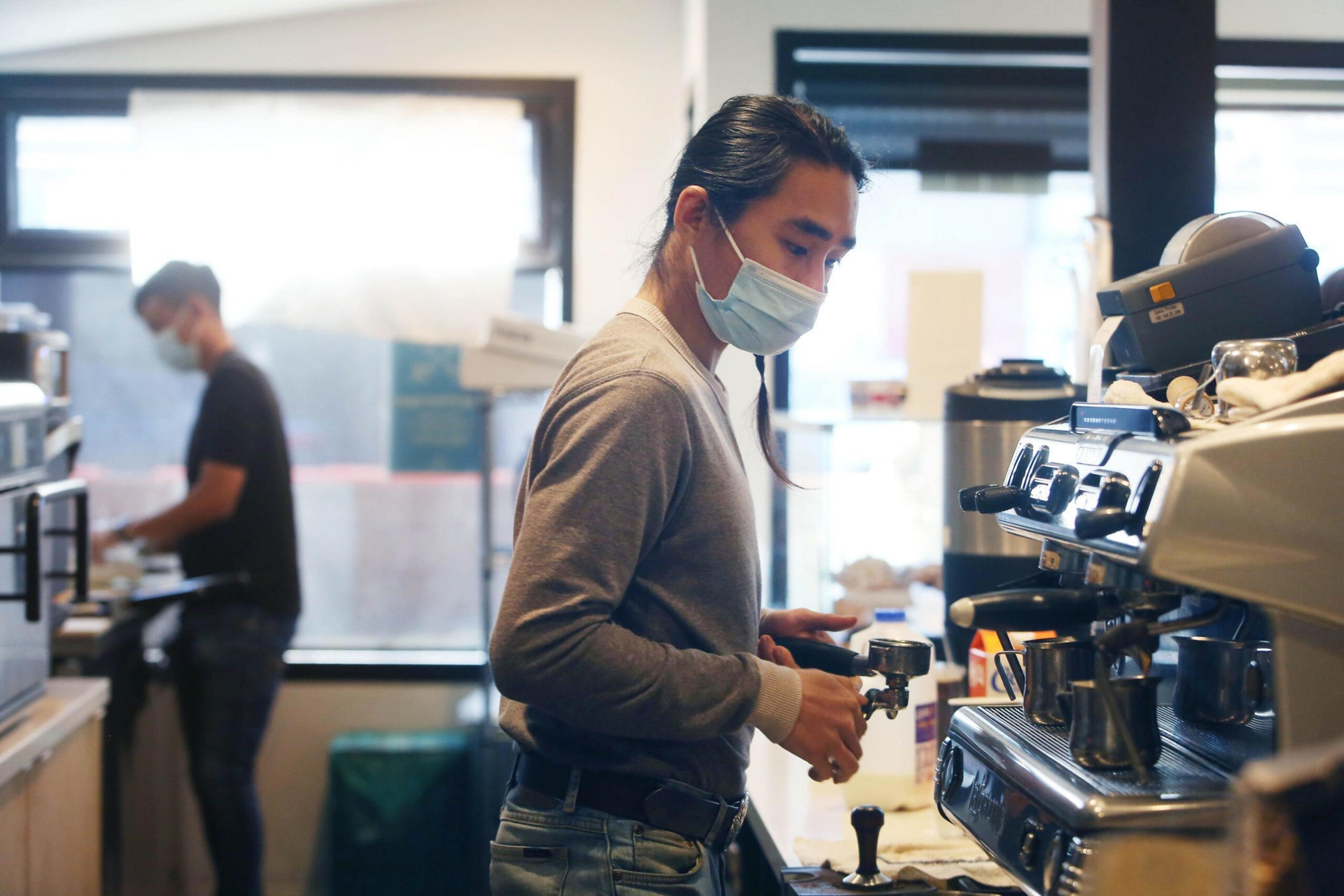 A masked barista prepares espresso using a machine while another masked worker operates in the background near a window in a coffee shop.
