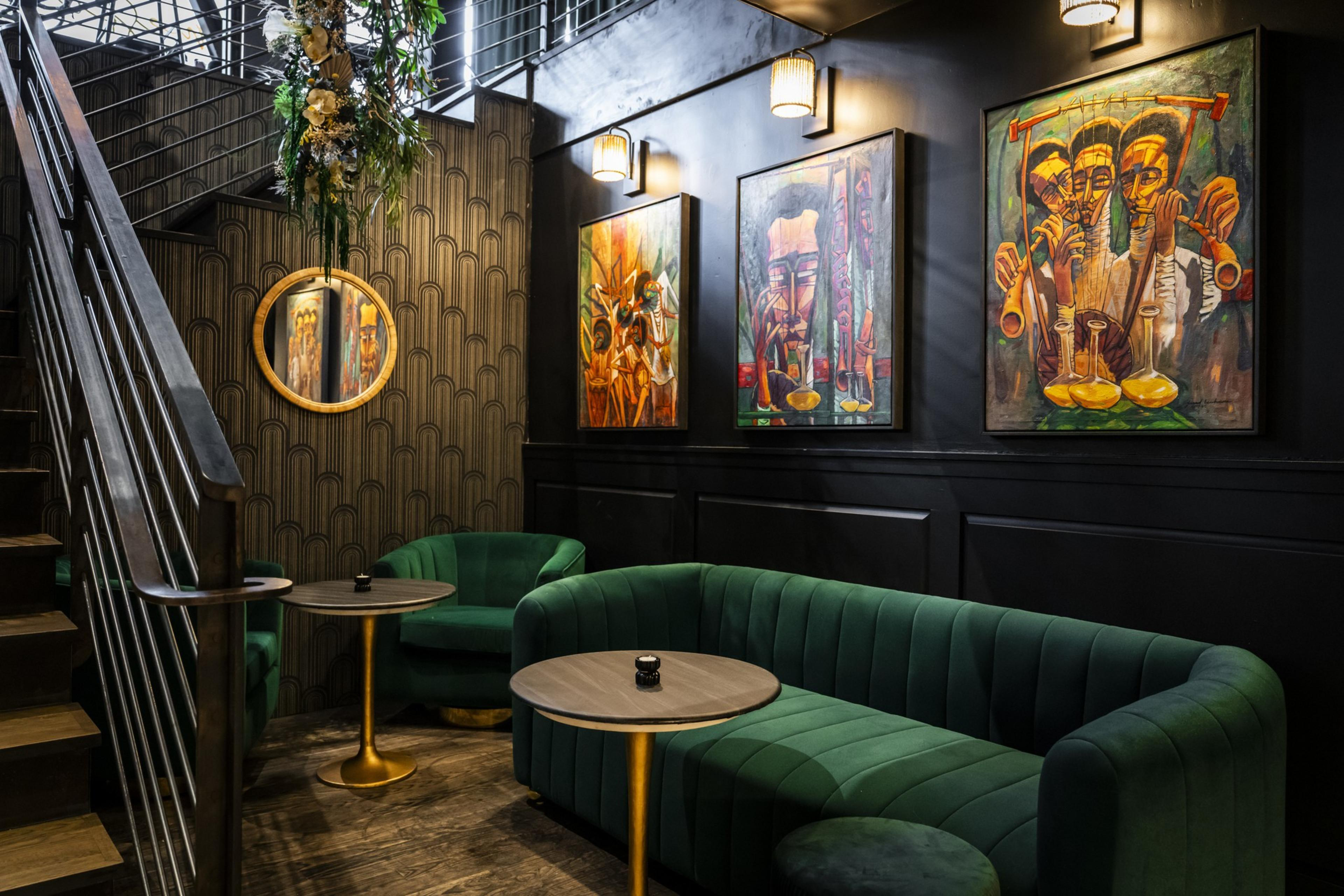 The image shows a cozy lounge with green velvet seating, gold round tables, and artistic paintings on dark walls. A circular mirror hangs on patterned wallpaper.