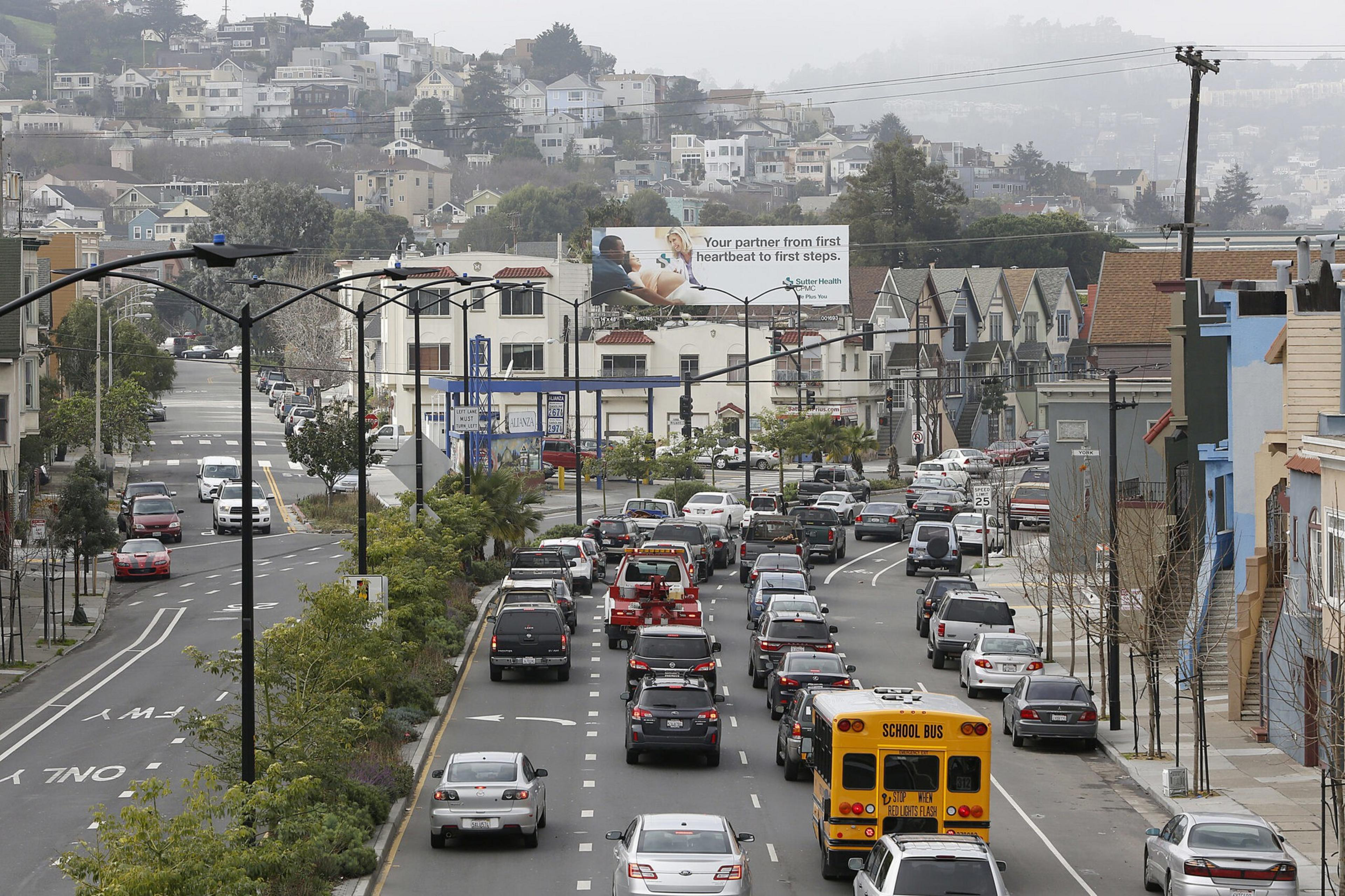 A city street with heavy traffic including cars and a yellow school bus, lined with houses and trees, set against a hillside with more houses in the background.