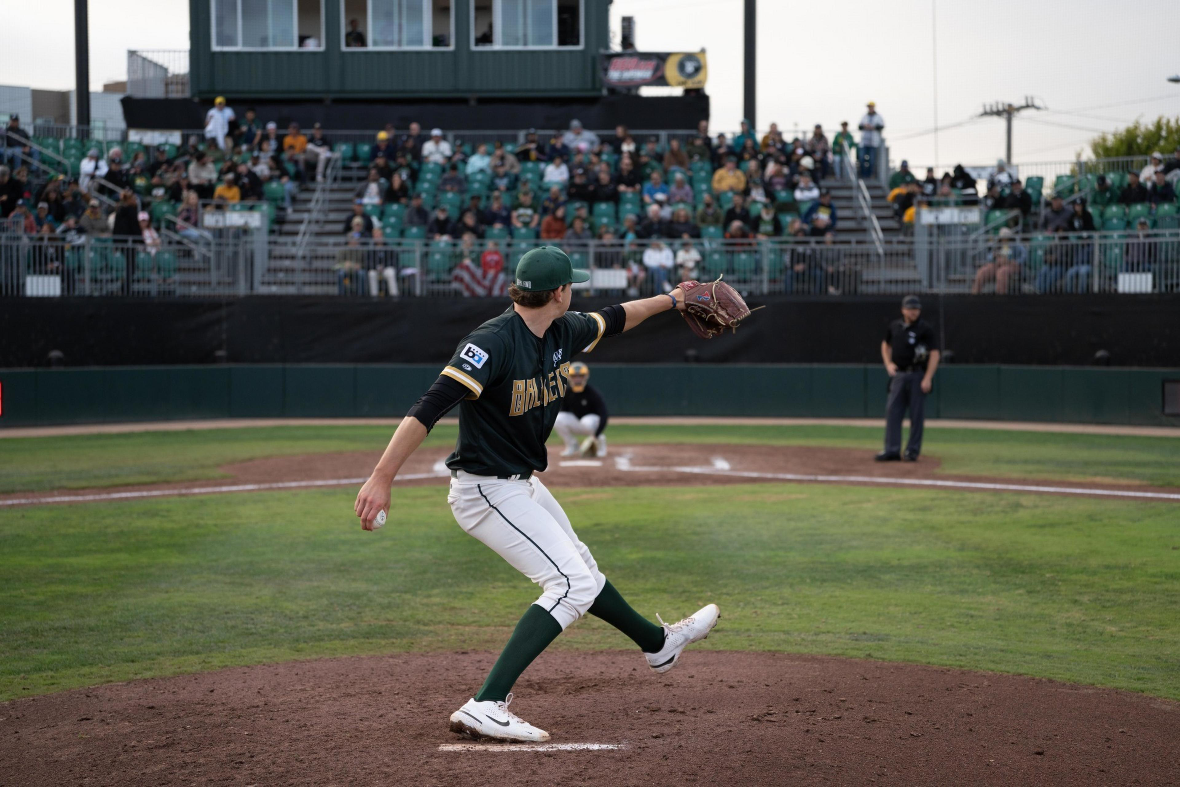 A baseball pitcher in green and white uniform is winding up to throw the ball with a catcher and umpire visible behind home plate and spectators in the stands.