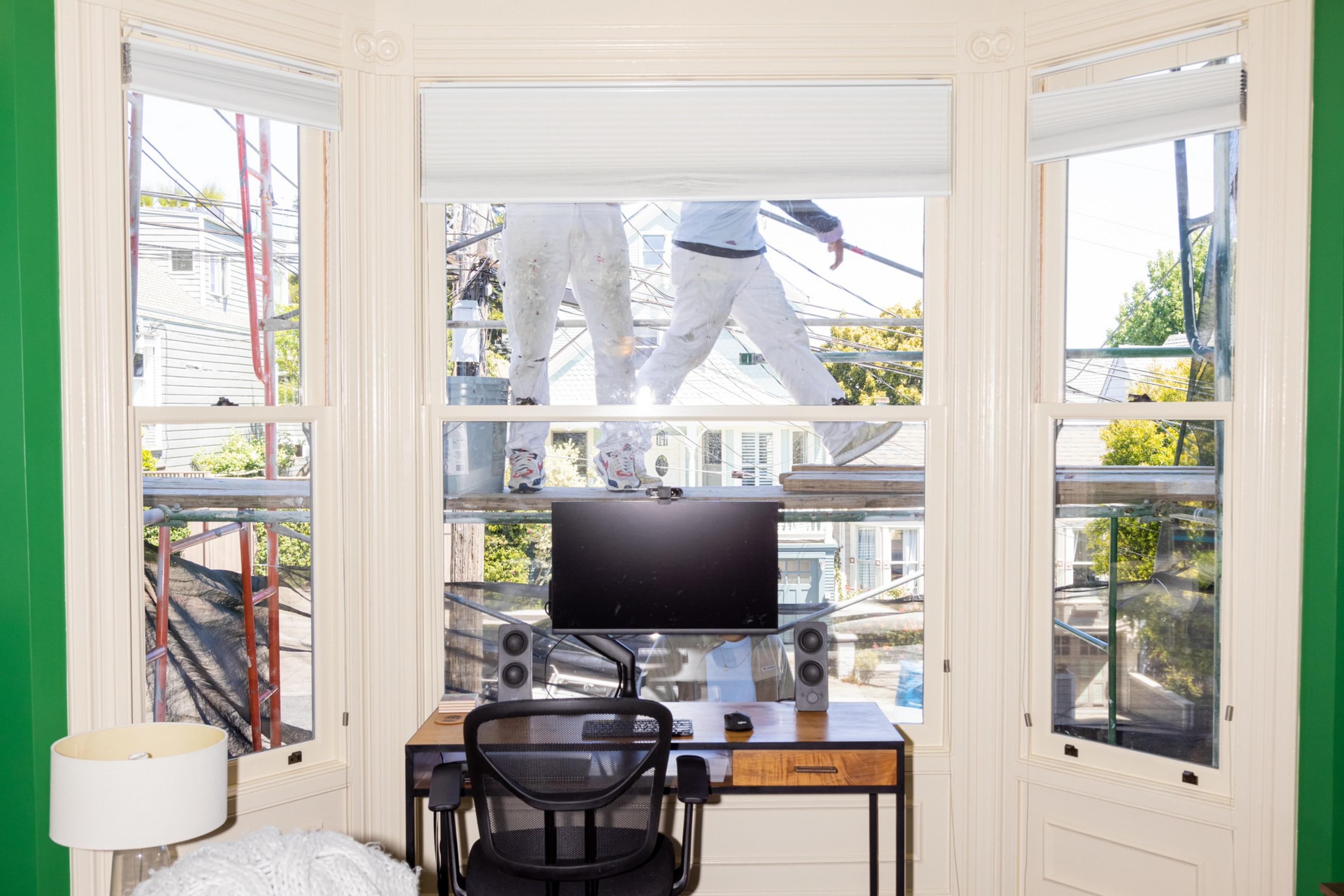 Two workers on scaffolding outside a window, viewed from an indoor room with a desk and computer.
