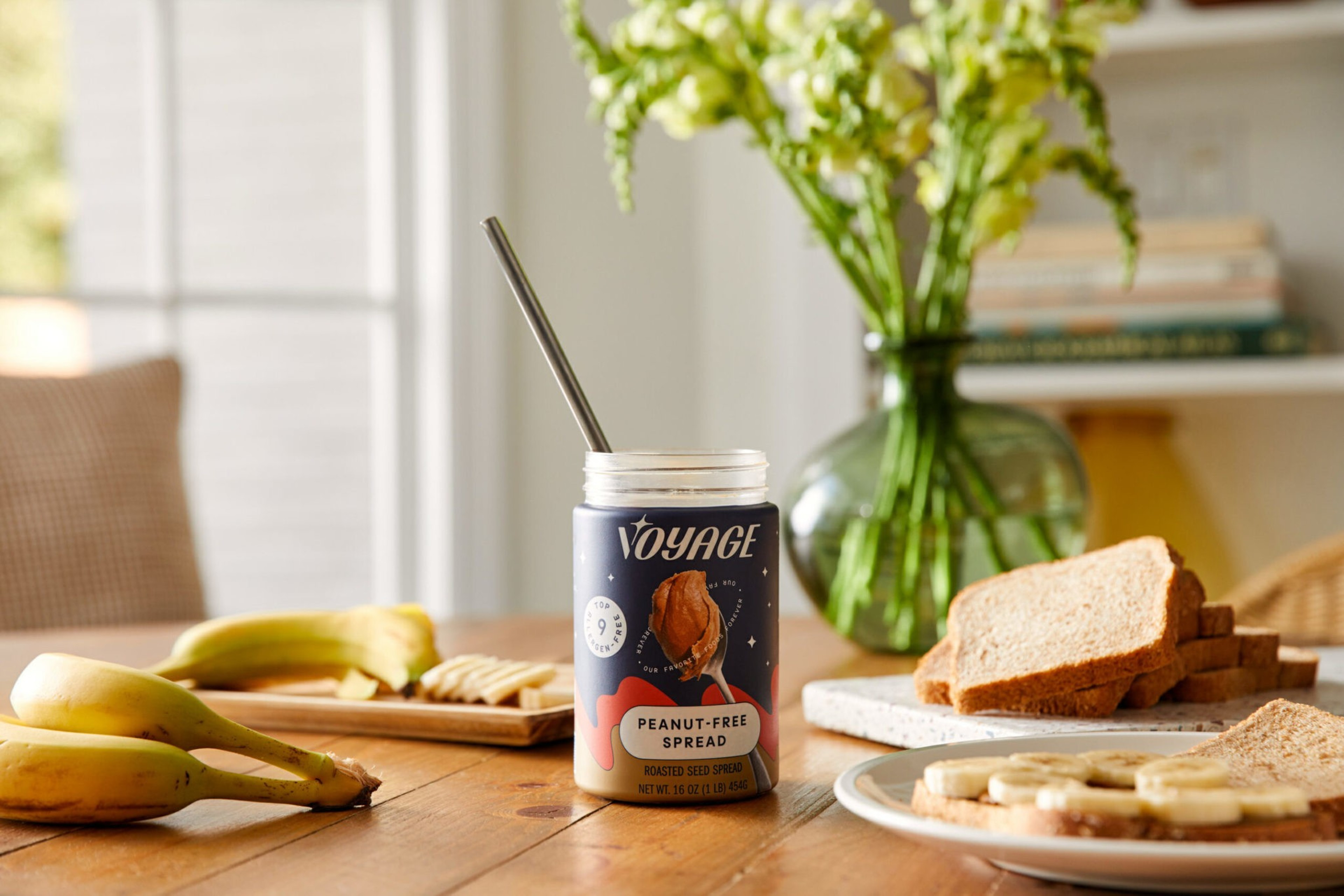 A jar of peanut-free roasted seed spread with a spoon sits on a table surrounded by bananas, crackers, bread slices, and a plate with banana slices.