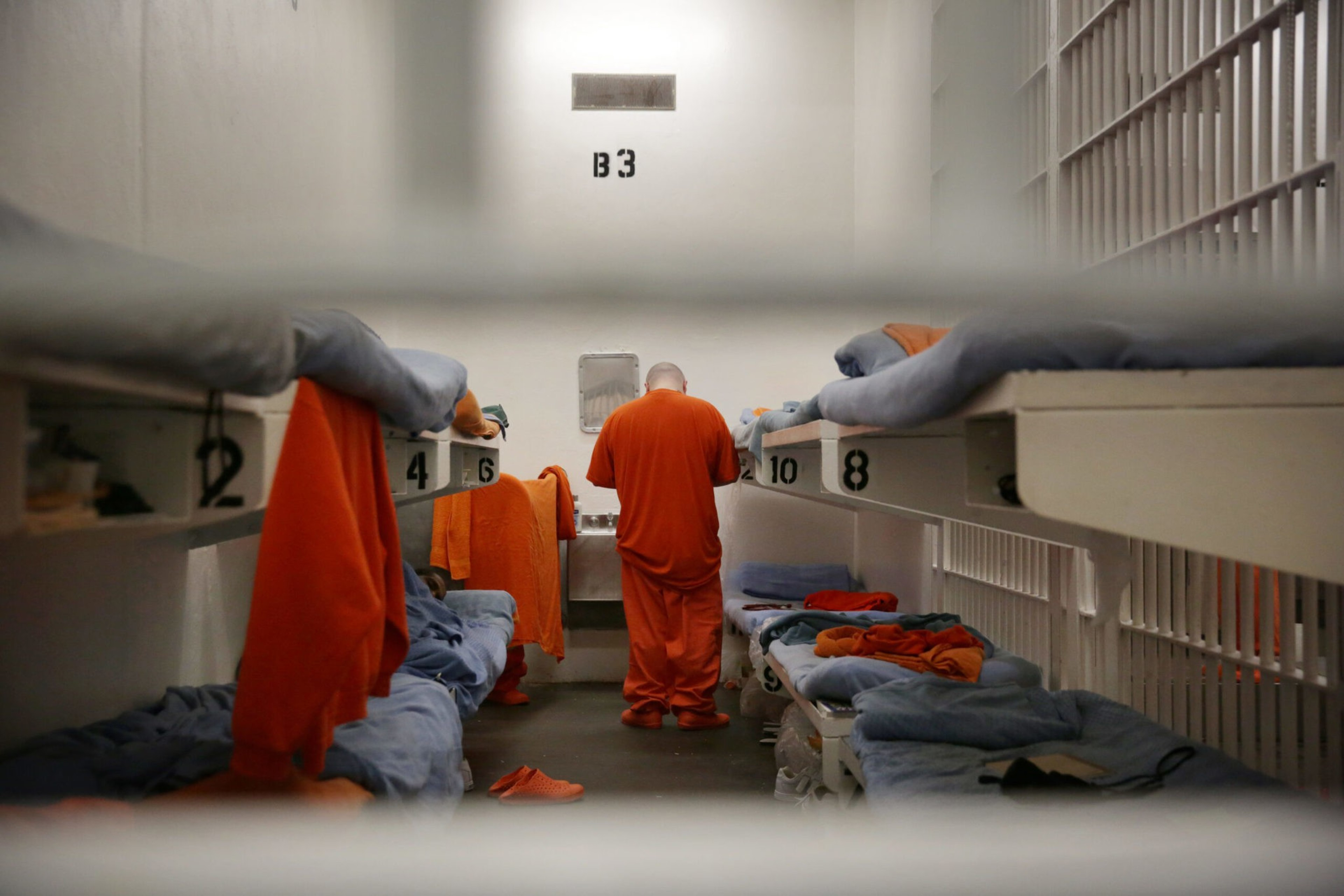 A prisoner in an orange jumpsuit stands with his back to the camera inside a small cell containing bunk beds with folded blue bedding and orange clothing.