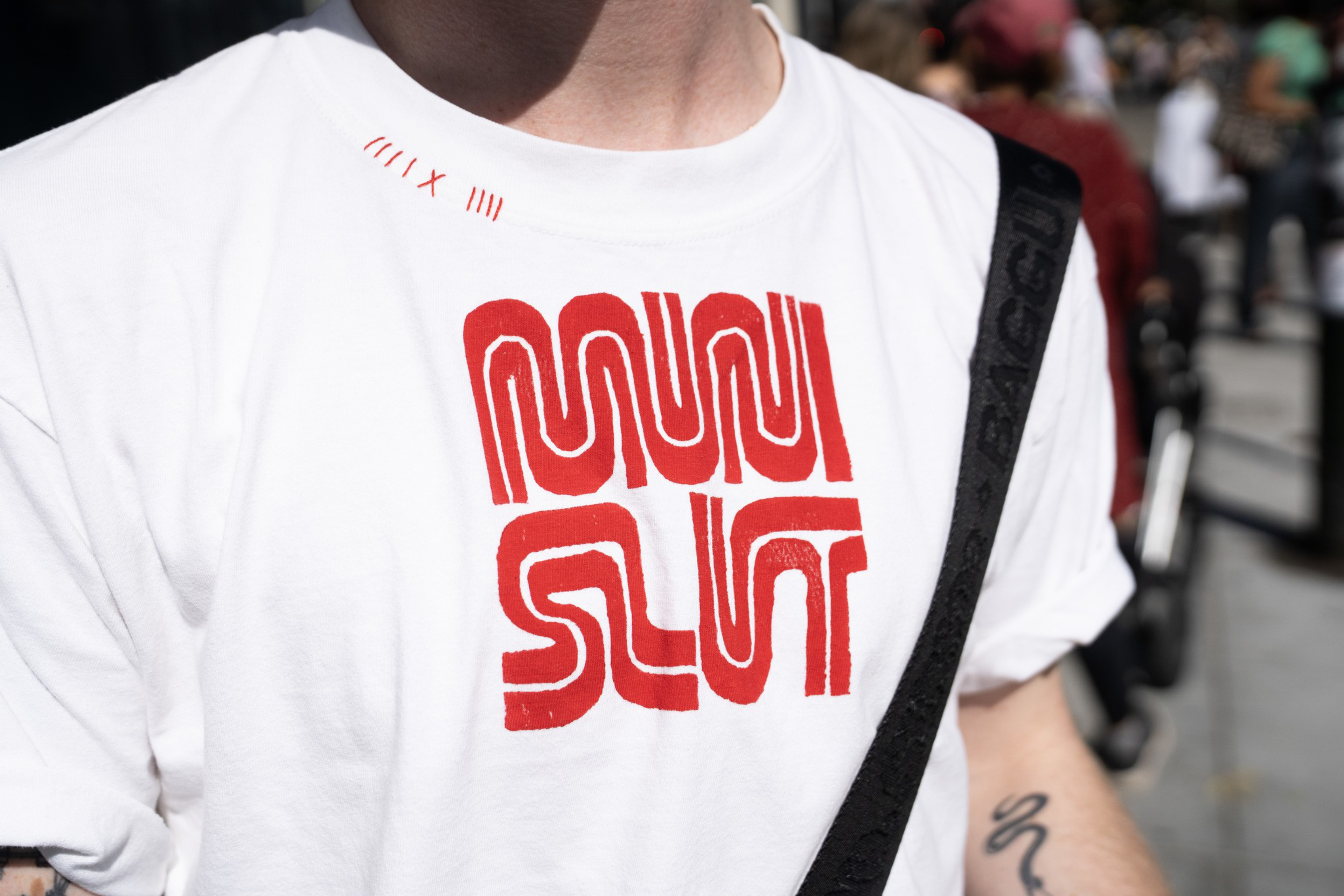 A person wears a white shirt with bold red letters spelling “MUNI SLUT” and small red Roman numerals near the collar.