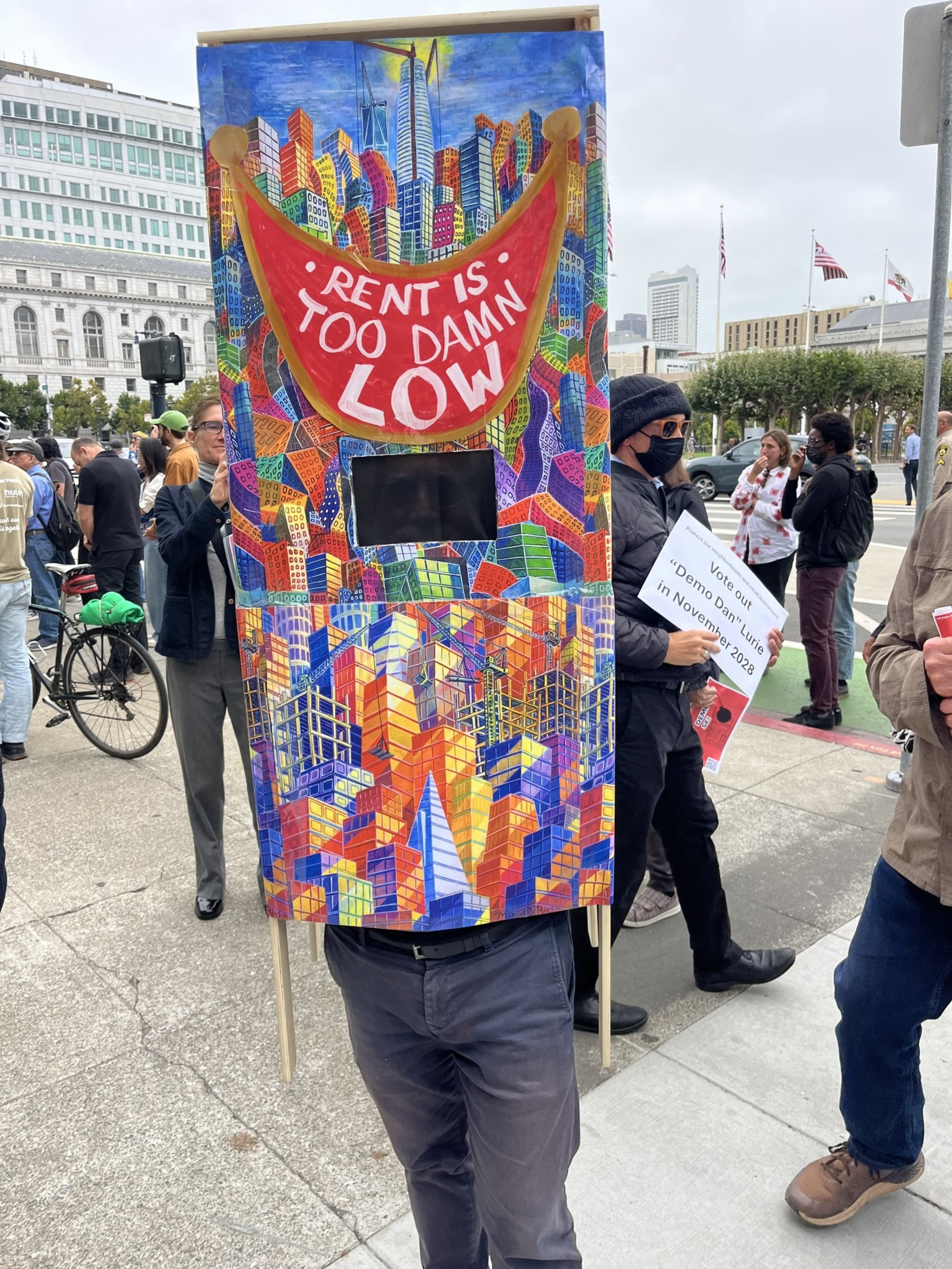 A person wears a colorful cityscape costume with a large red banner reading "RENT IS TOO DAMN LOW" at a street protest with other people nearby.