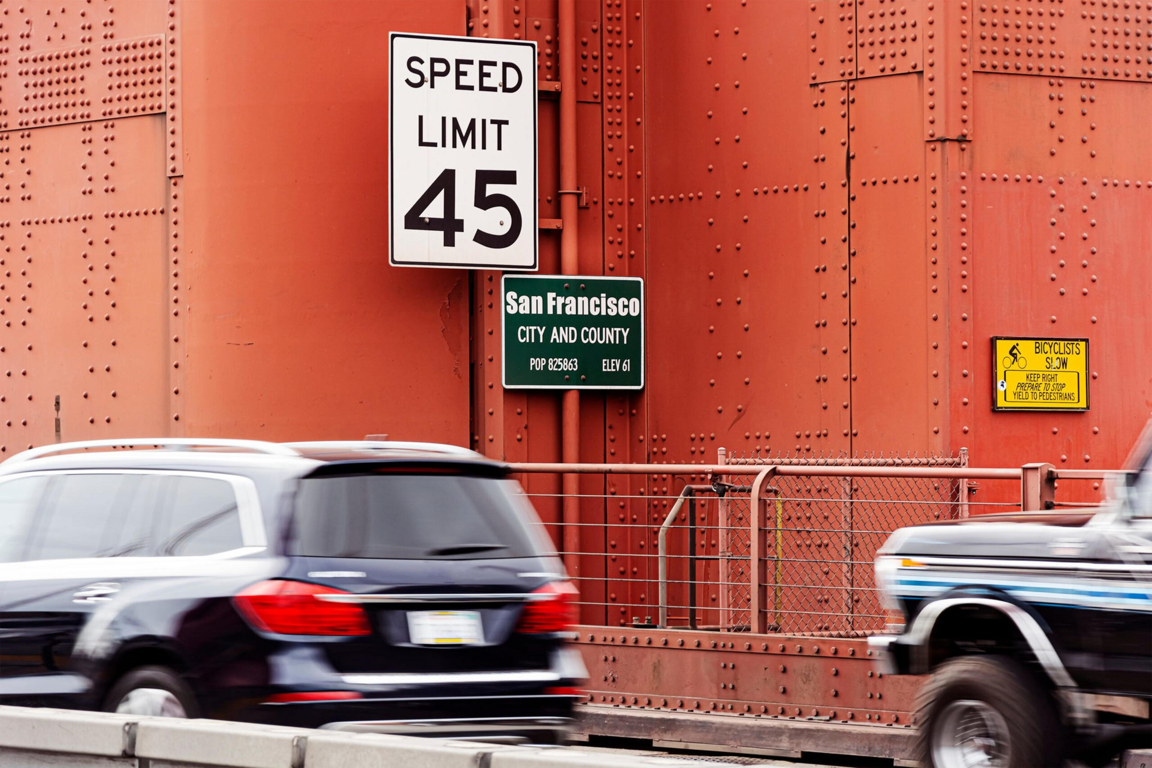 Two vehicles pass a red metal structure with signs indicating a 45 mph speed limit, San Francisco city and county, and bicyclists should keep right.