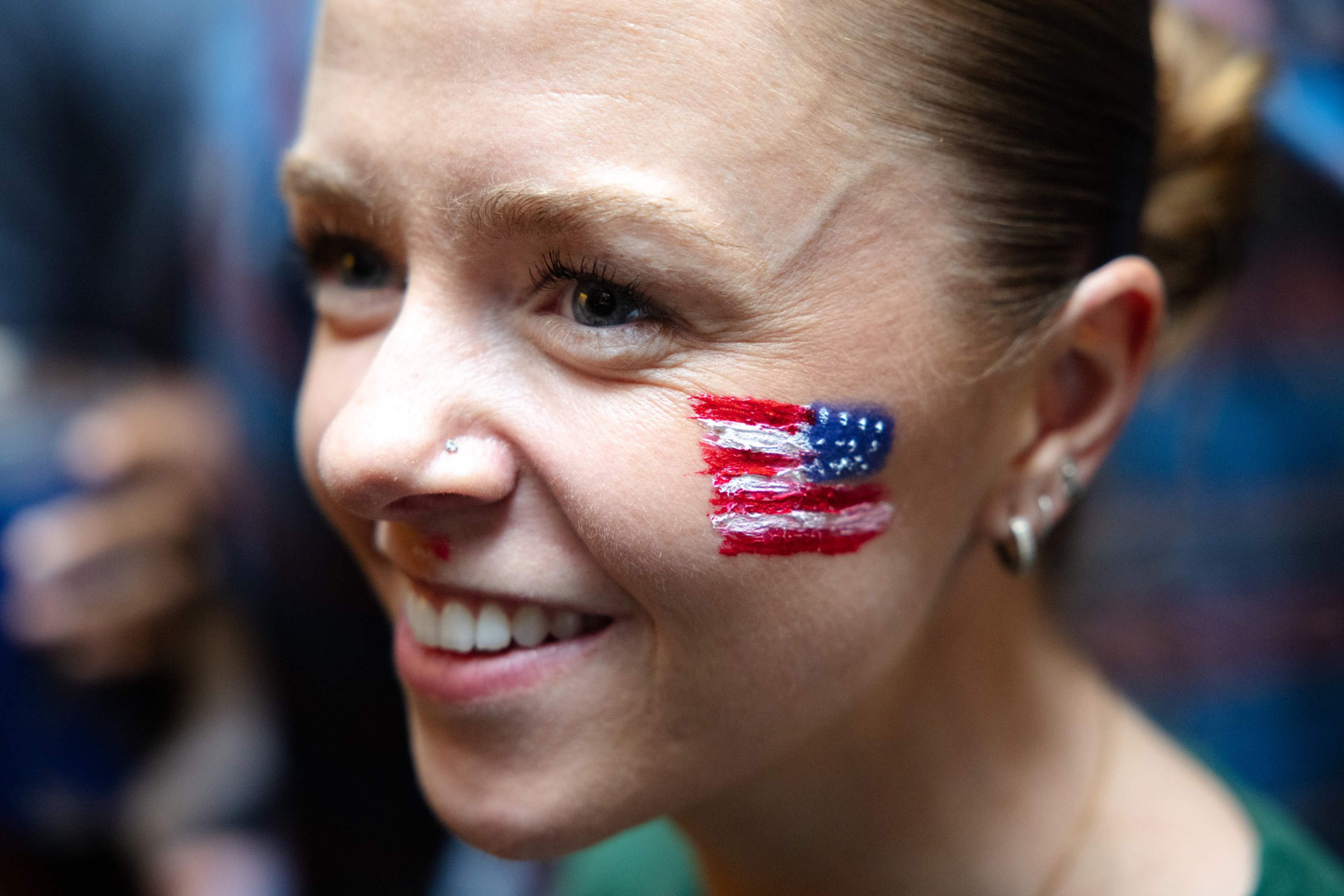 A smiling woman with blonde hair has a painted American flag on her right cheek and wears a small nose ring and hoop earrings.