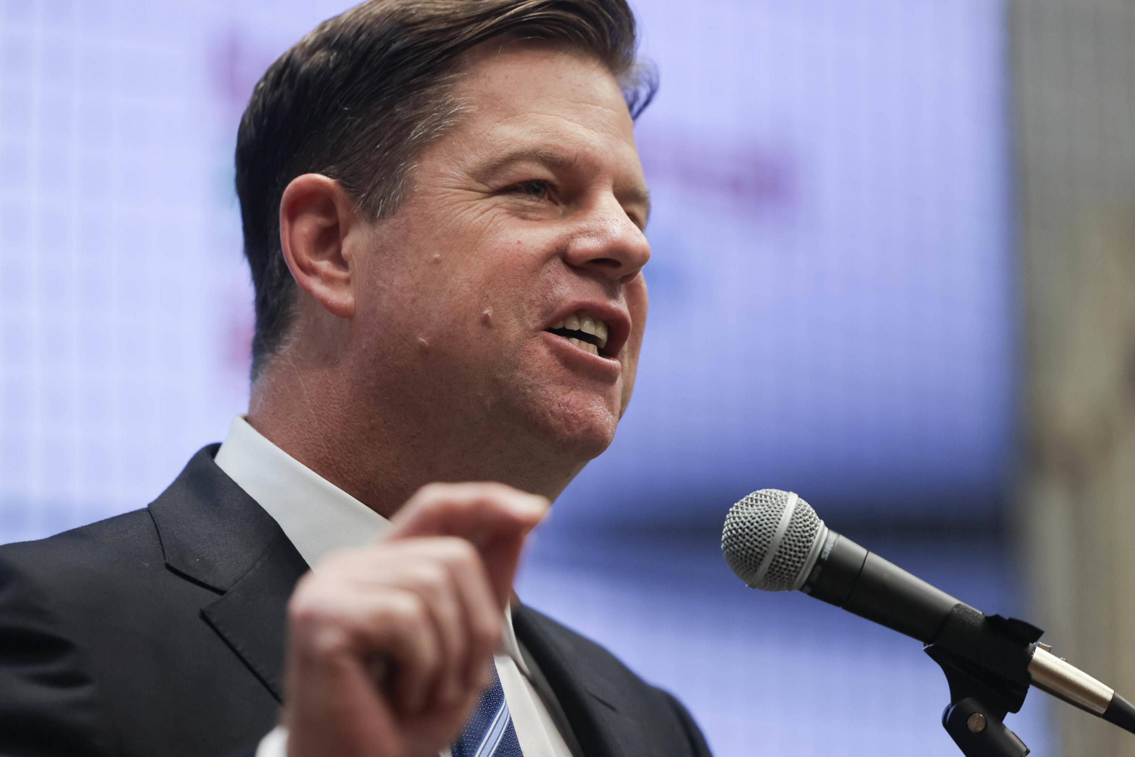 A man in a suit speaks in front of a microphone pointing with his finger.