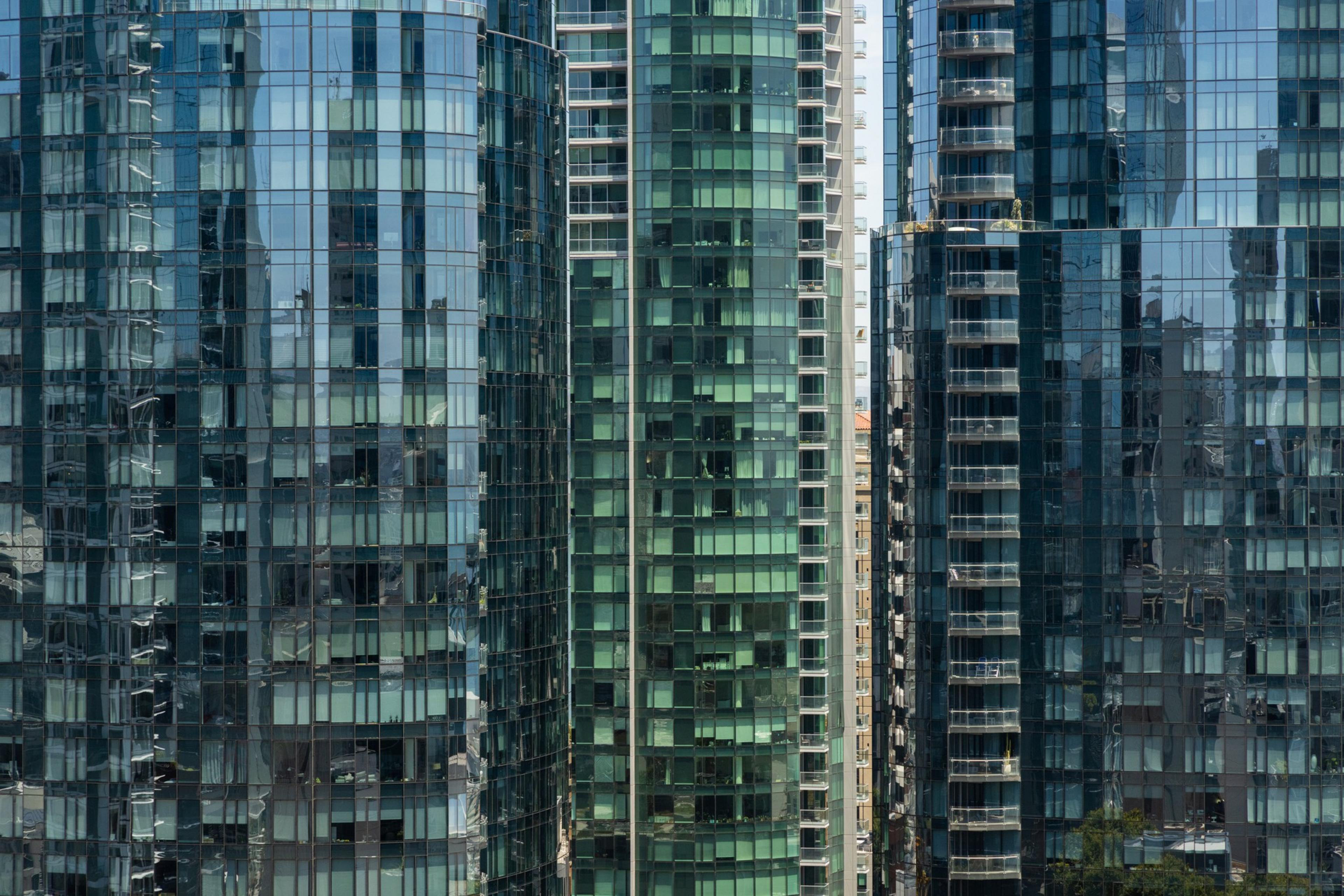 The image shows several tall glass skyscrapers closely packed together, reflecting the sky and neighboring buildings. The buildings have a modern, sleek design.