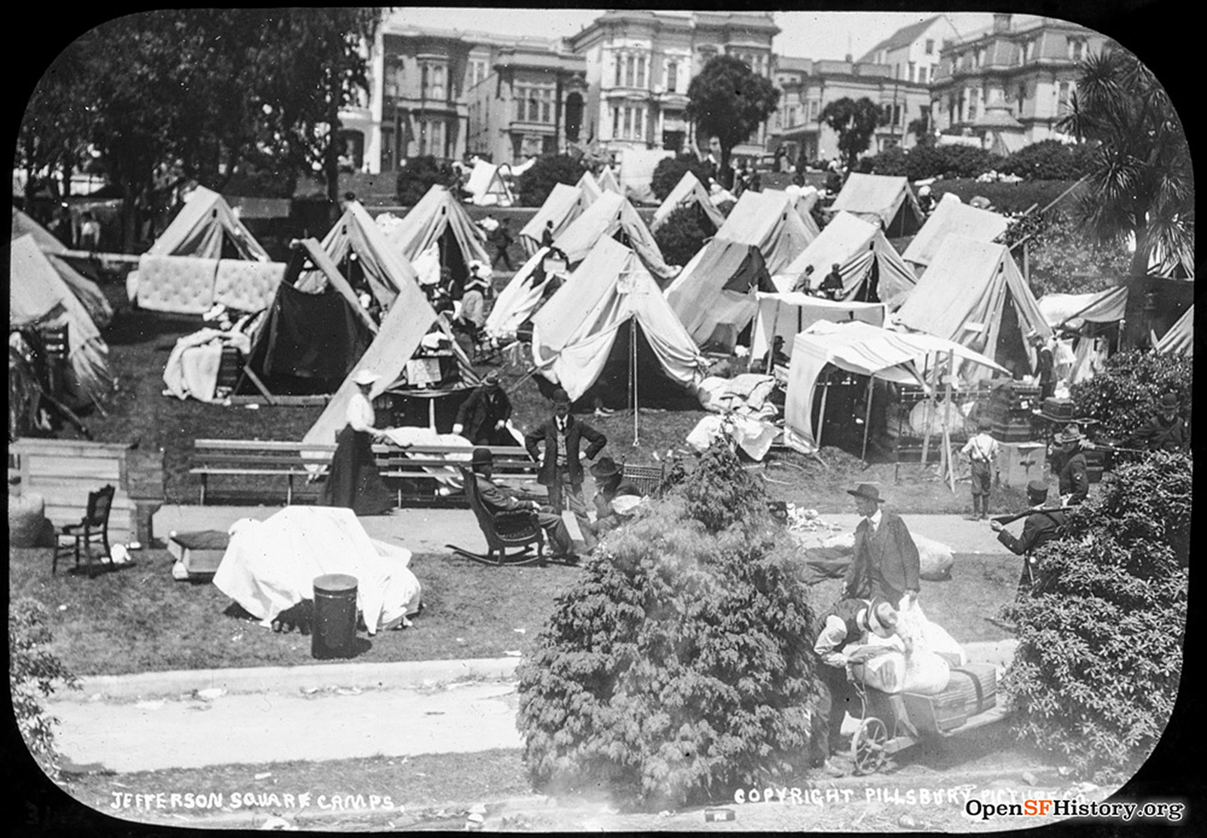 Rows of canvas tents set up in a park with several people walking and sitting among them, surrounded by ornate buildings in the background.
