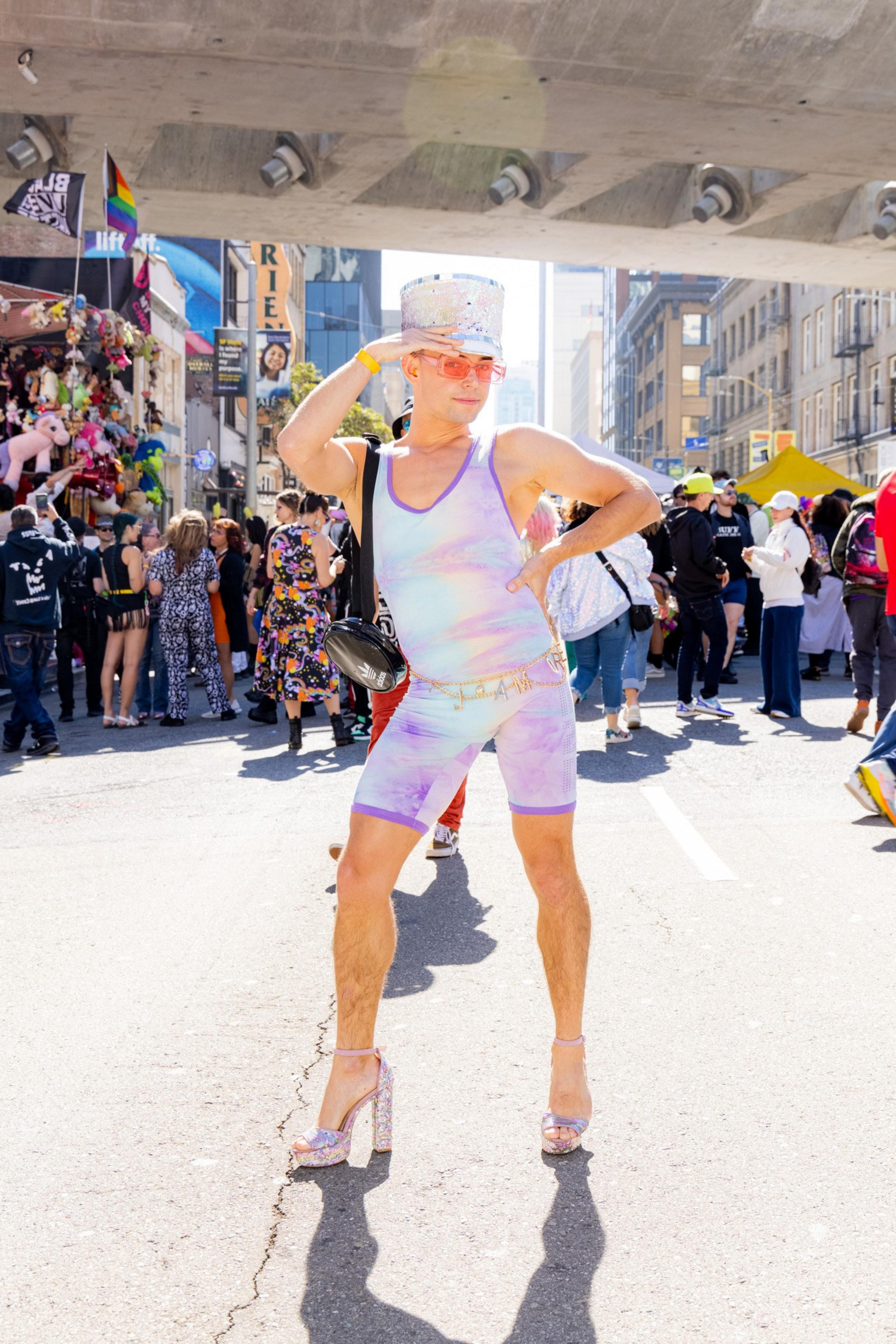 A person dressed in a pastel rainbow bodysuit and sparkling high heels strikes a pose on a street. They wear a glittery top hat and sunglasses, with a lively crowd behind them.