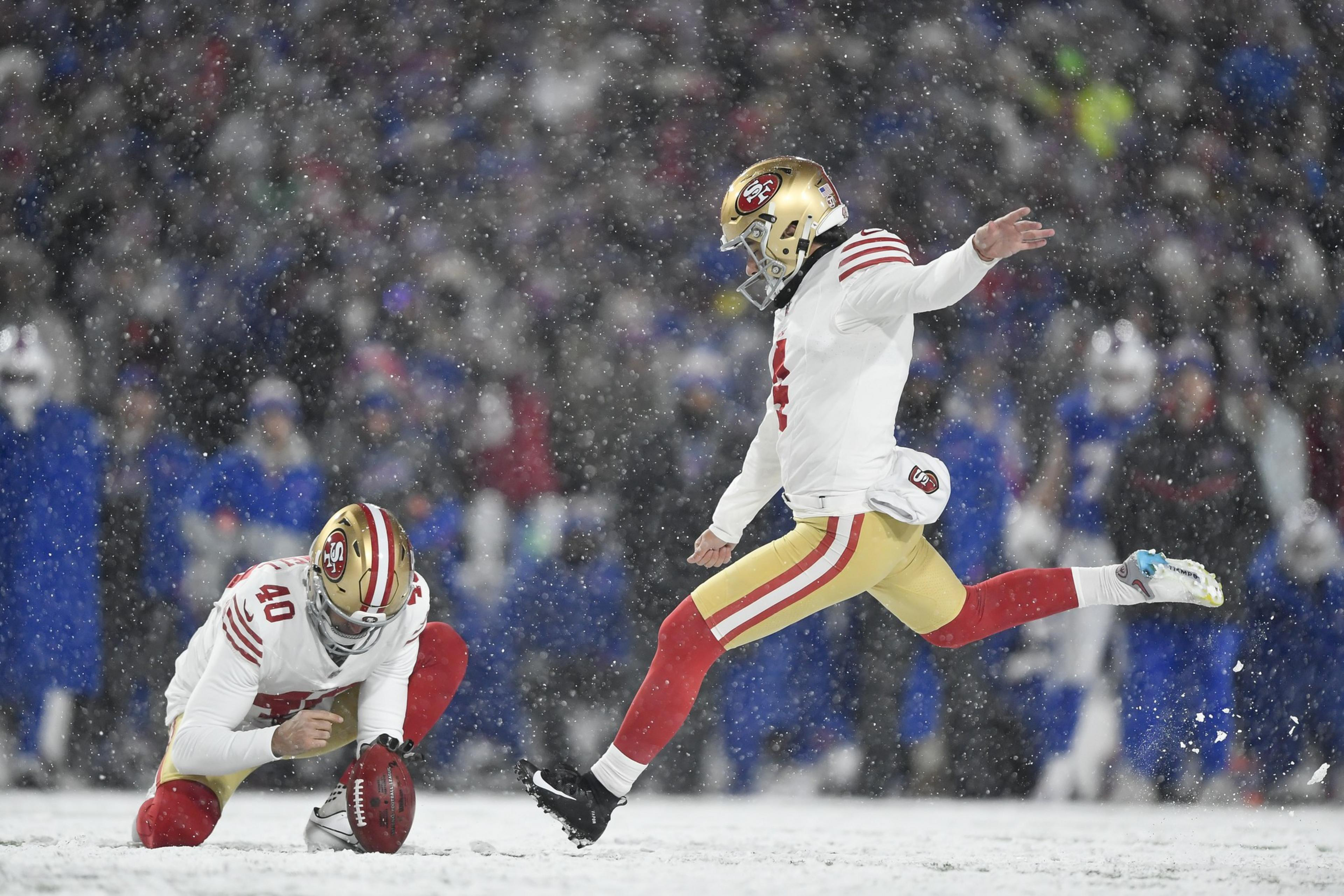 A football player in a snowy field kicks a ball held by a teammate. They're wearing white jerseys with red and gold pants, and snow falls around them.