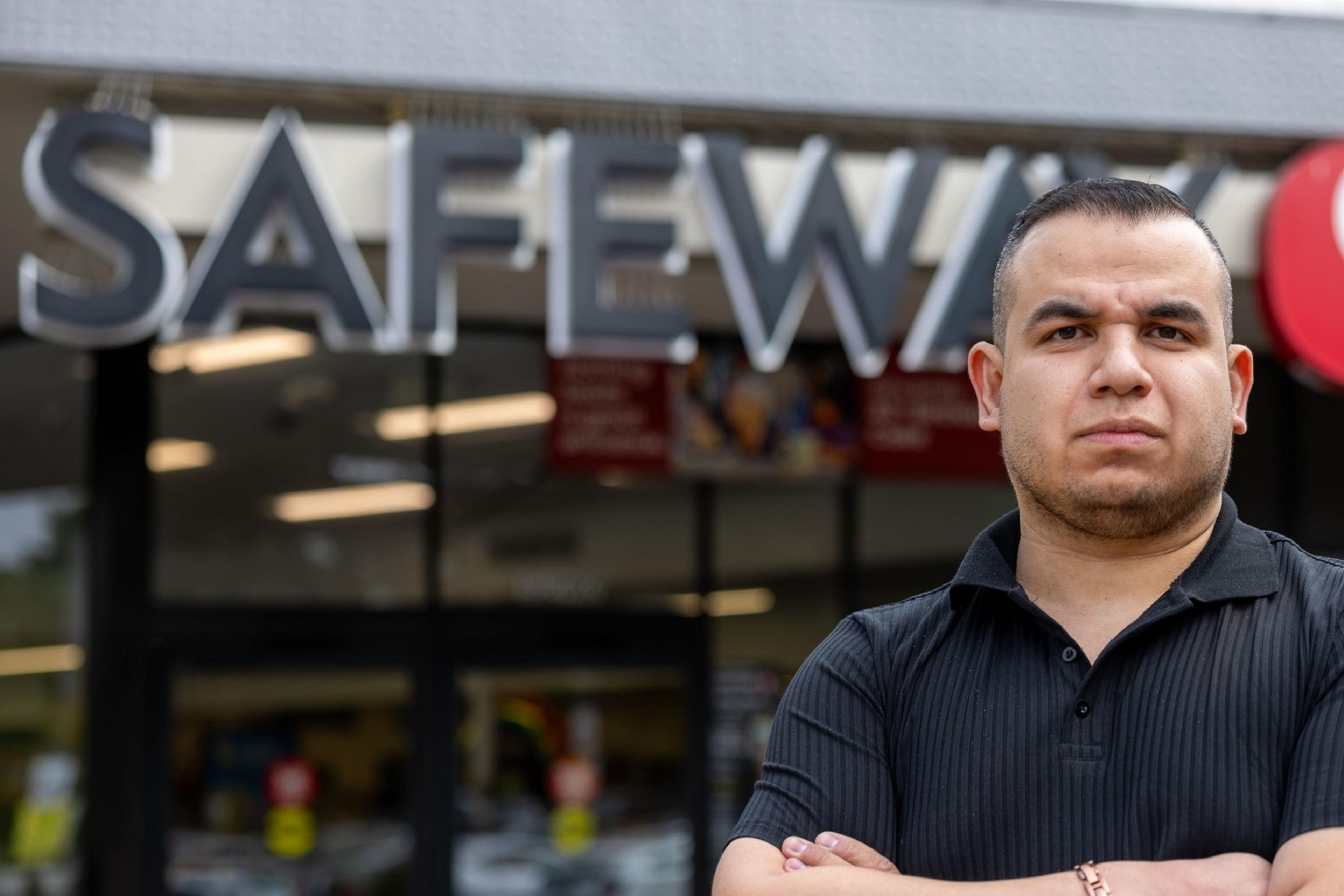 A man stands with arms crossed in front of a Safeway supermarket. He is wearing a black polo shirt and appears serious, with the store's signage clearly visible above him.