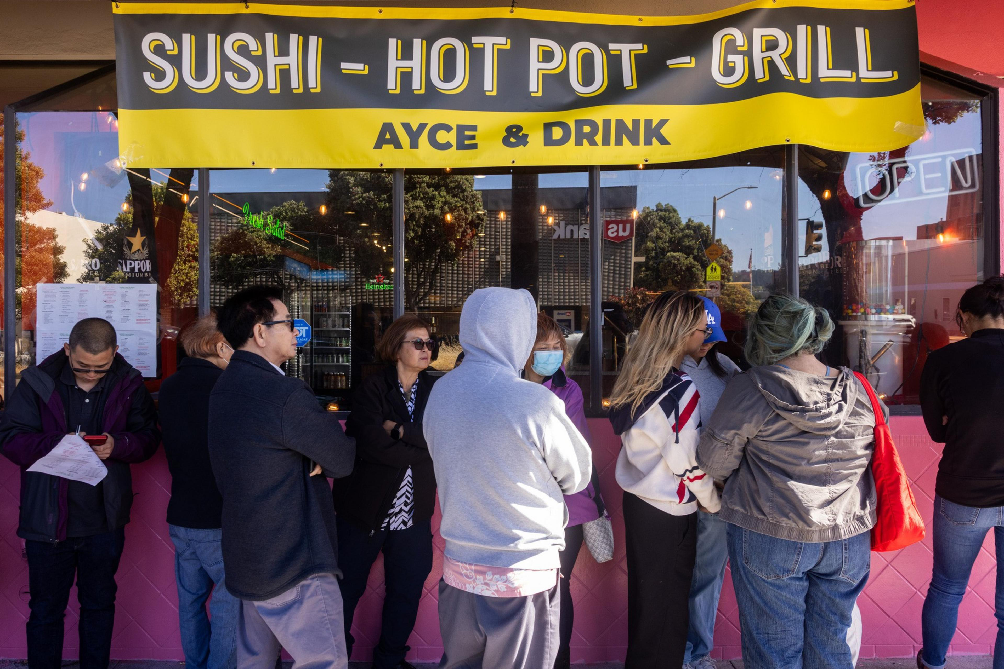 People stand in line outside a restaurant with a bright yellow sign reading "Sushi - Hot Pot - Grill, AYCE & Drink." The scene is lively and social.