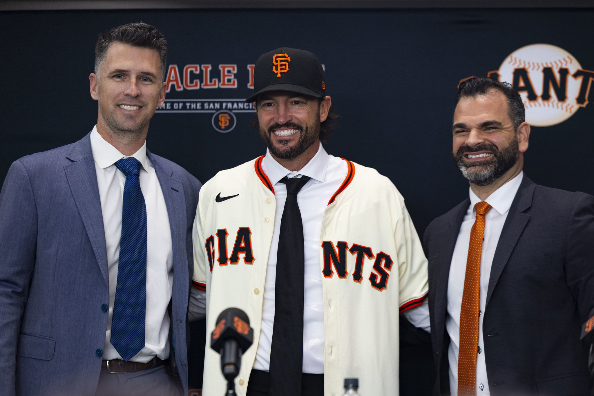 Three men stand smiling; the center man wears a San Francisco Giants jersey and cap, while the other two wear suits and ties.