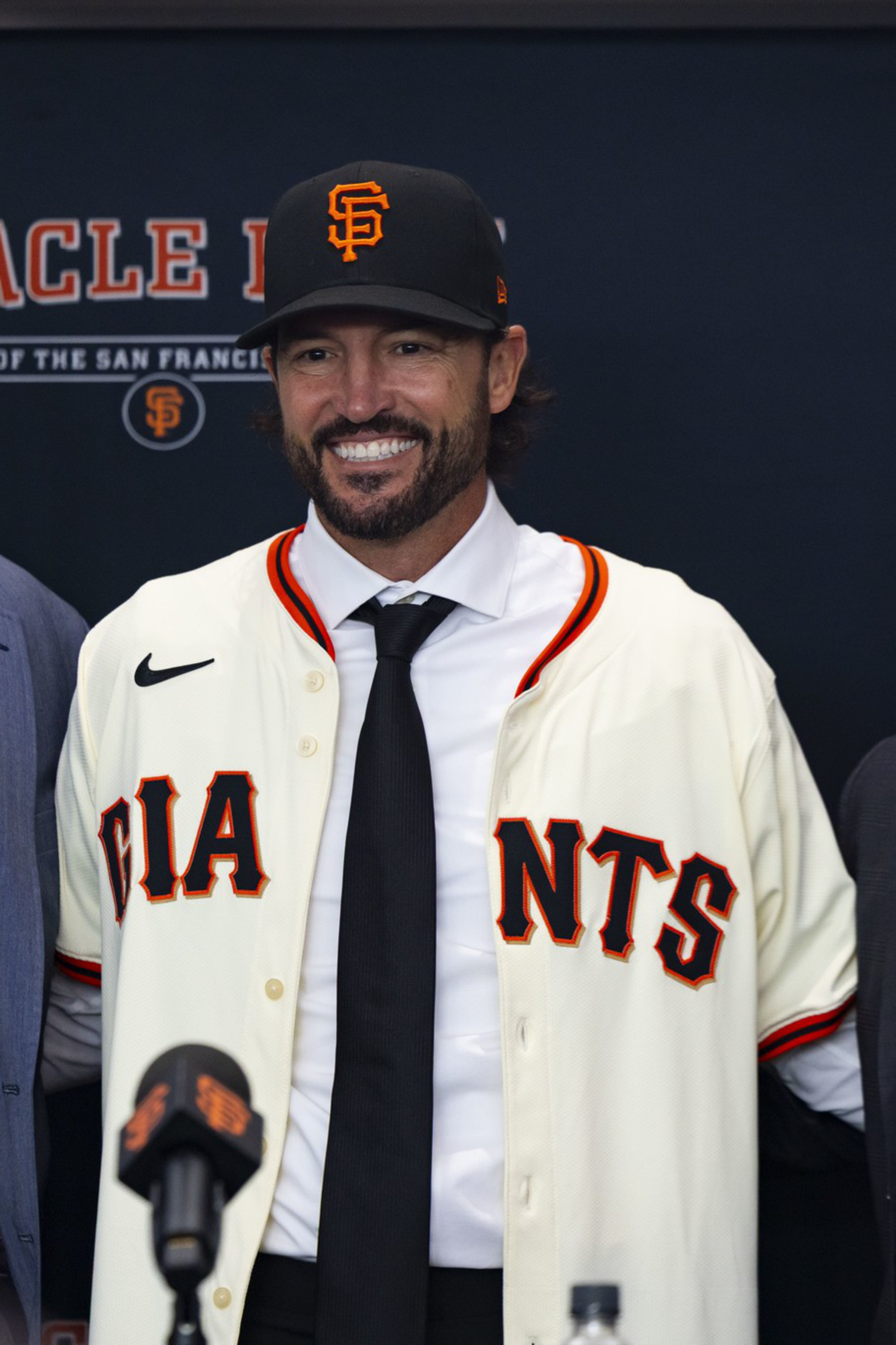 Three men stand smiling; the center man wears a San Francisco Giants jersey and cap, while the other two wear suits and ties.
