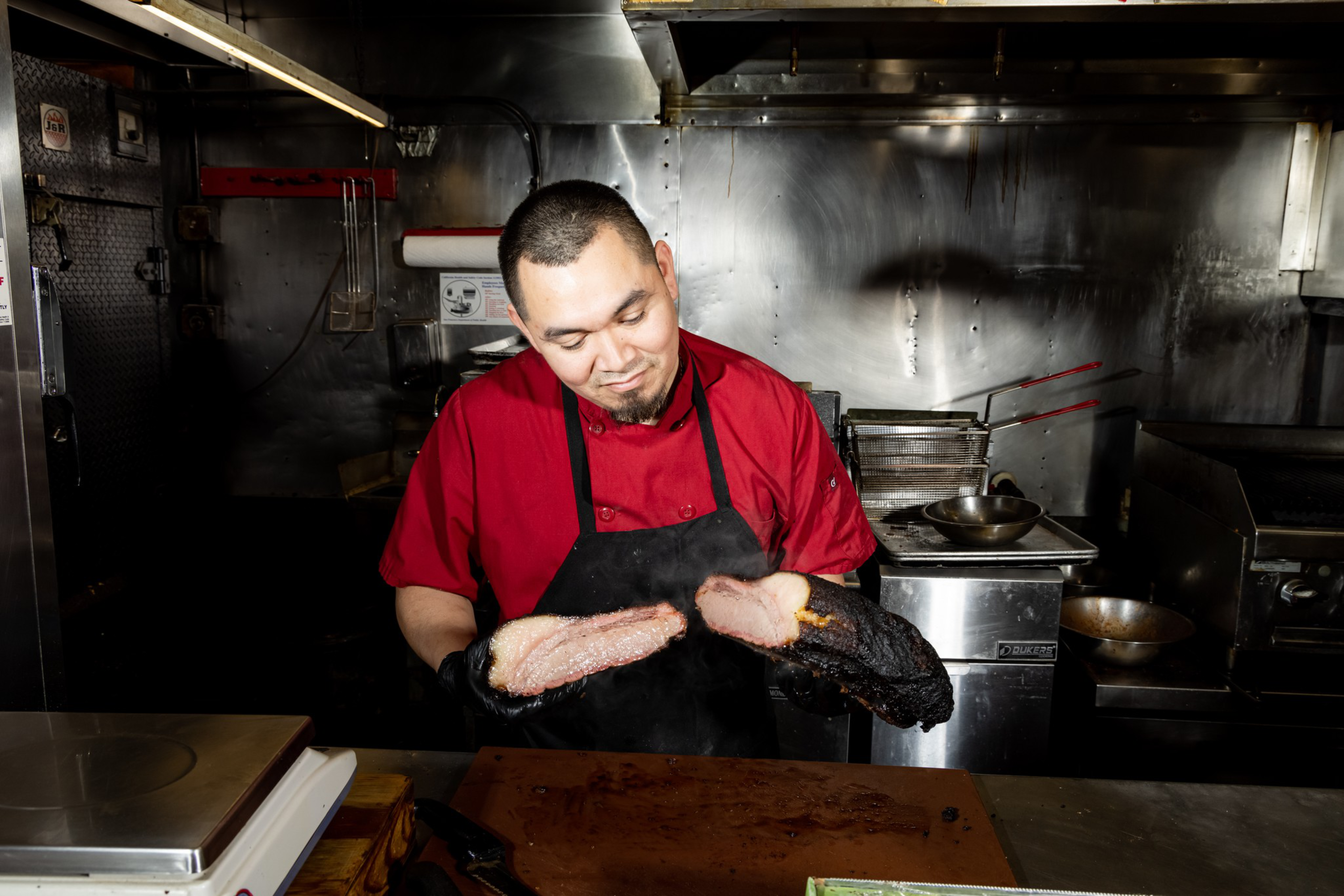 A chef in a red shirt and black apron holds two thick slices of smoked meat, one looking tender and the other charred, in a commercial kitchen.
