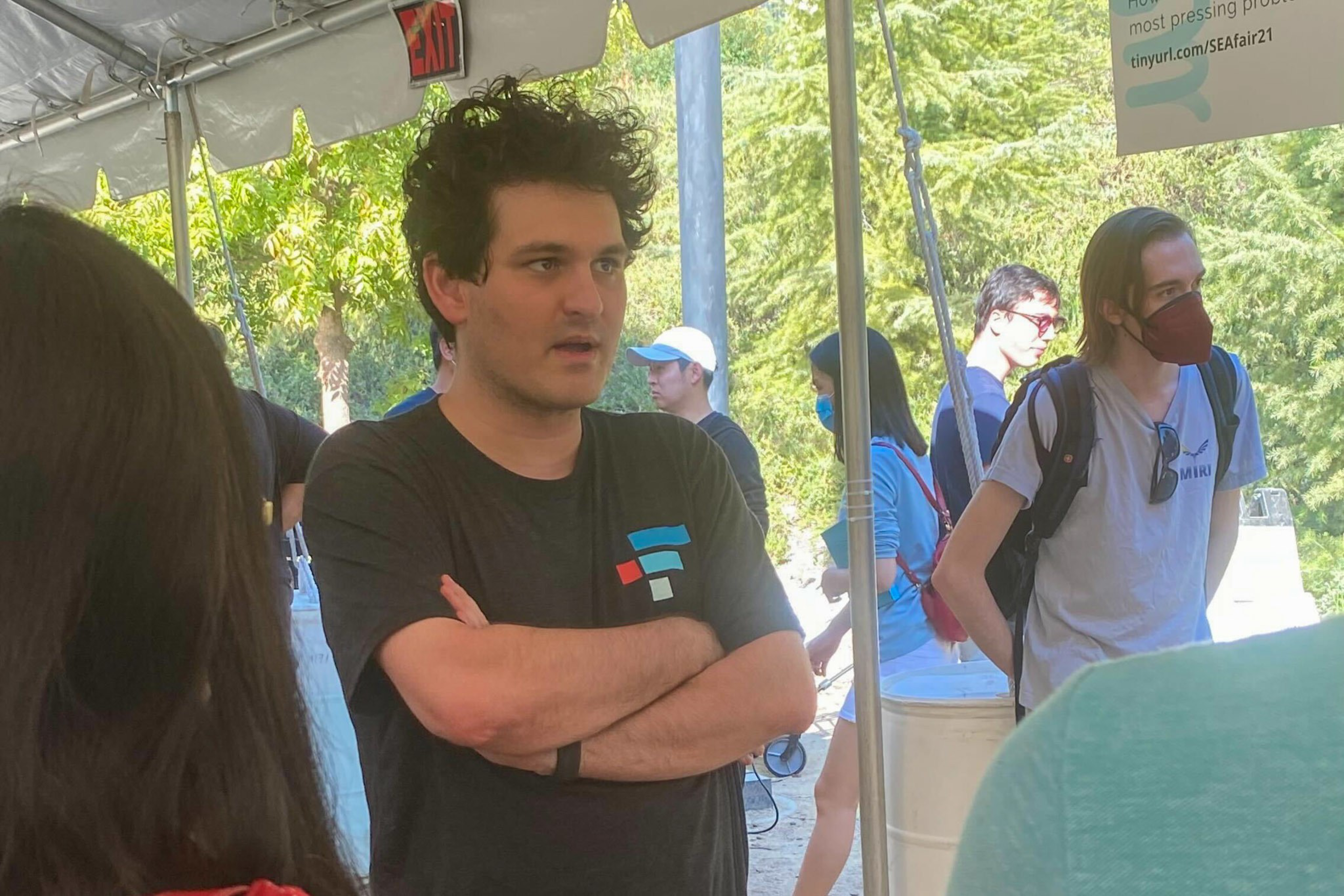 A man with curly hair and a dark T-shirt stands with arms crossed under a tent, while several people, some wearing masks, are nearby outside in a sunny area.