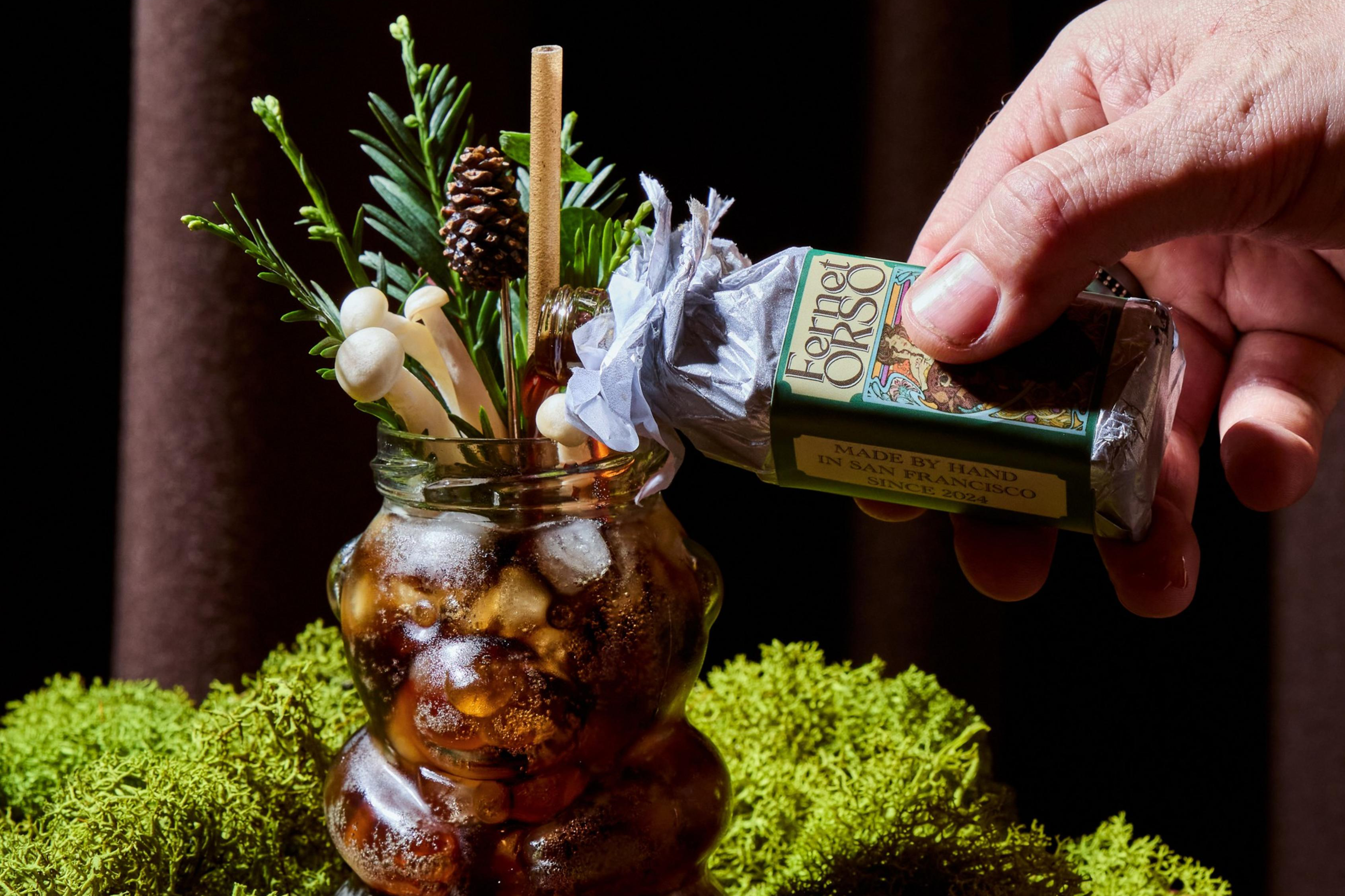 A hand sprays Fernet Orso from a small bottle into a bear-shaped glass filled with iced dark liquid and garnished with pine, mushrooms, and a pine cone.