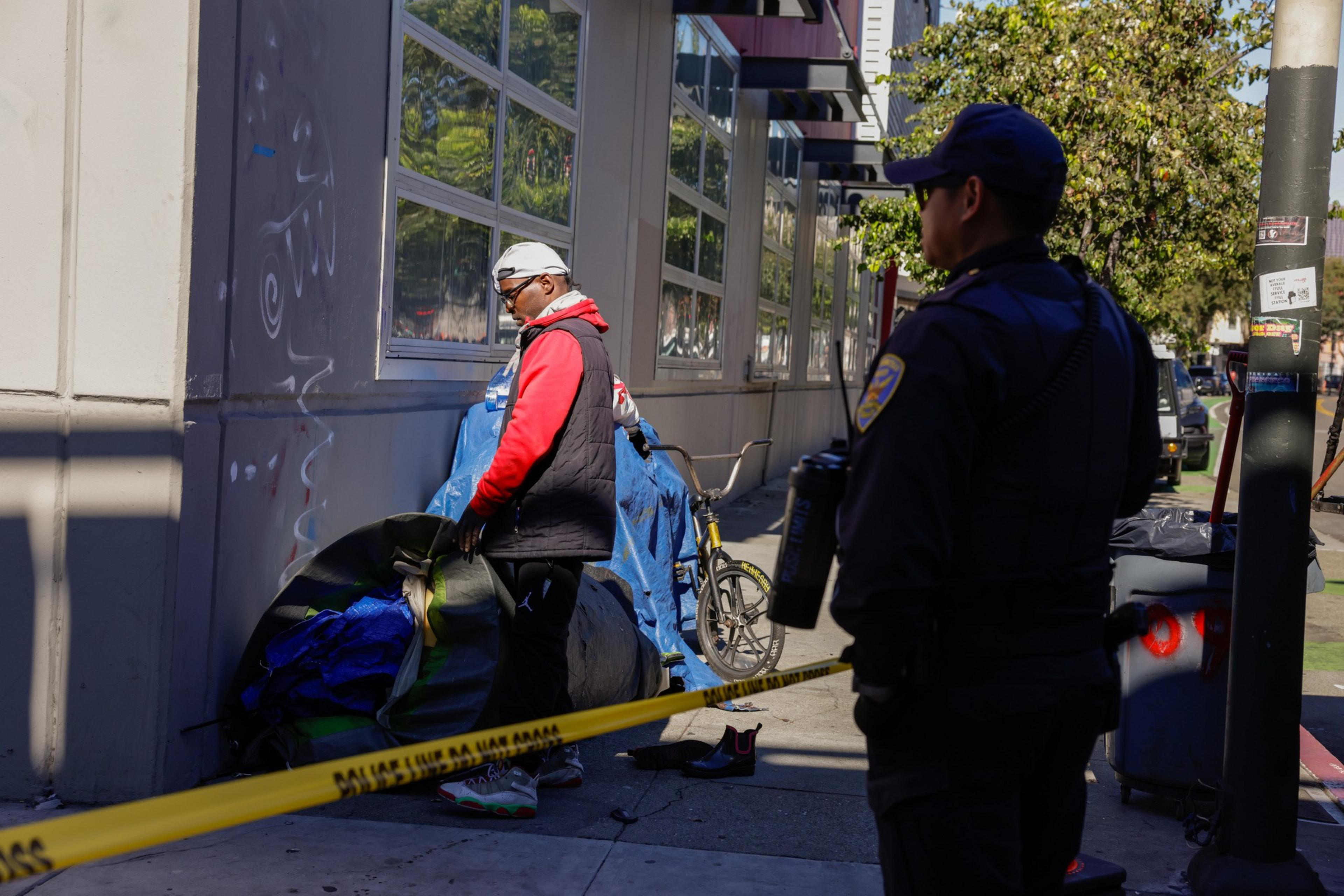 A man stands near a wall with a bicycle and belongings covered in a tarp. A police officer observes, and caution tape partially blocks the area.