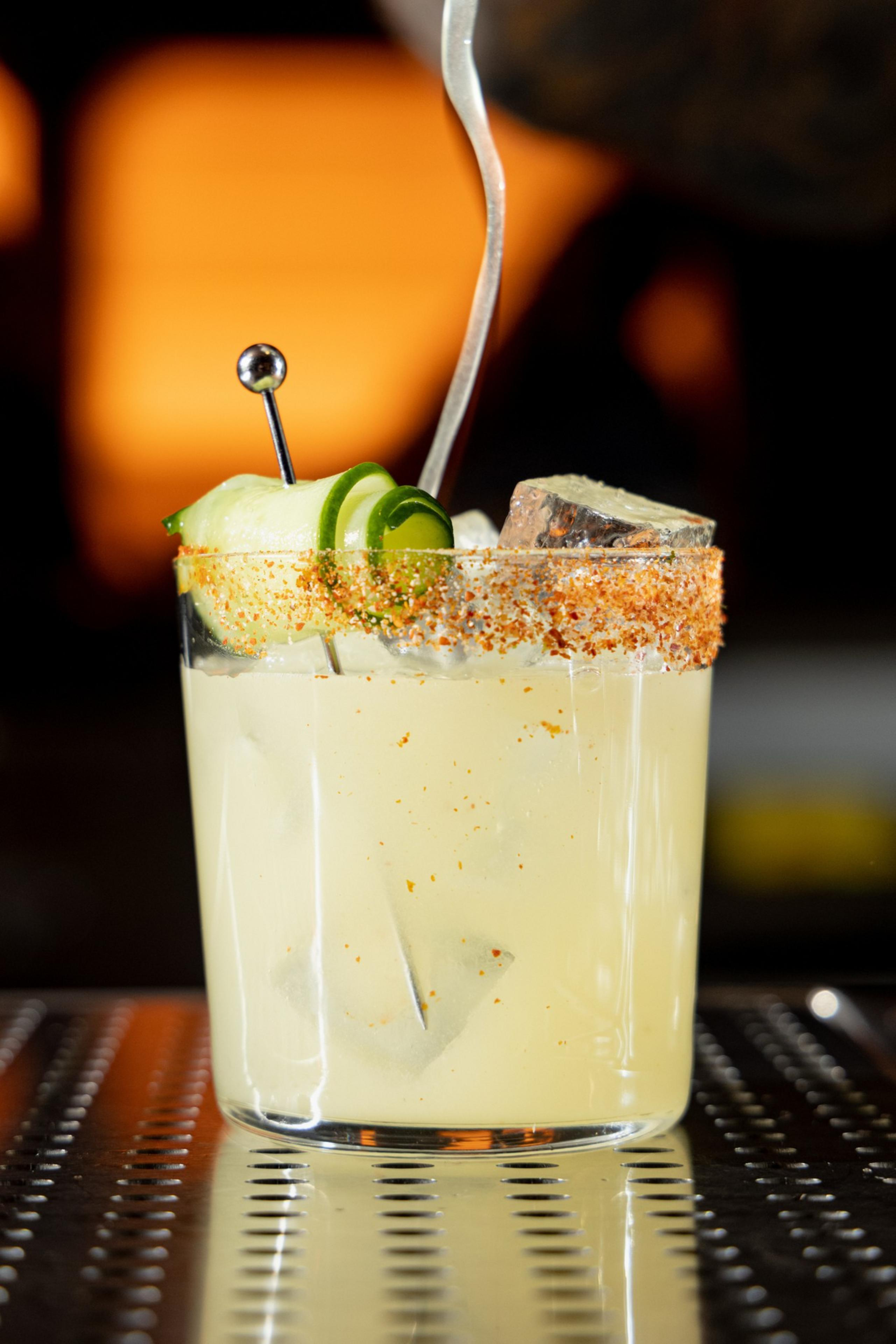 A pale yellow cocktail in a glass with chili powder rim, ice cubes, cucumber slices on a skewer, and a metal spoon inside.