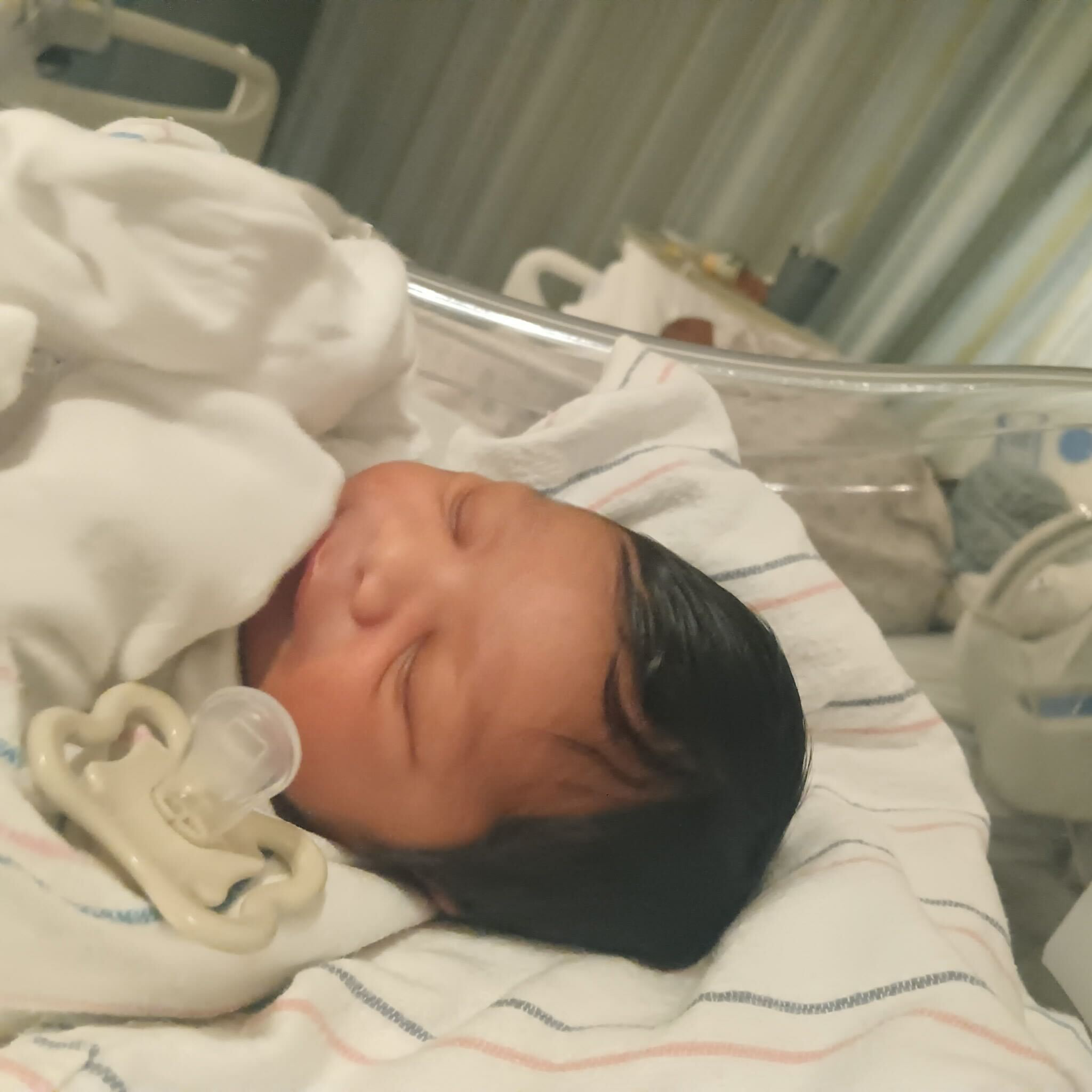 A newborn baby with dark hair is swaddled in a hospital blanket, lying in a clear hospital bassinet with a pacifier beside them.