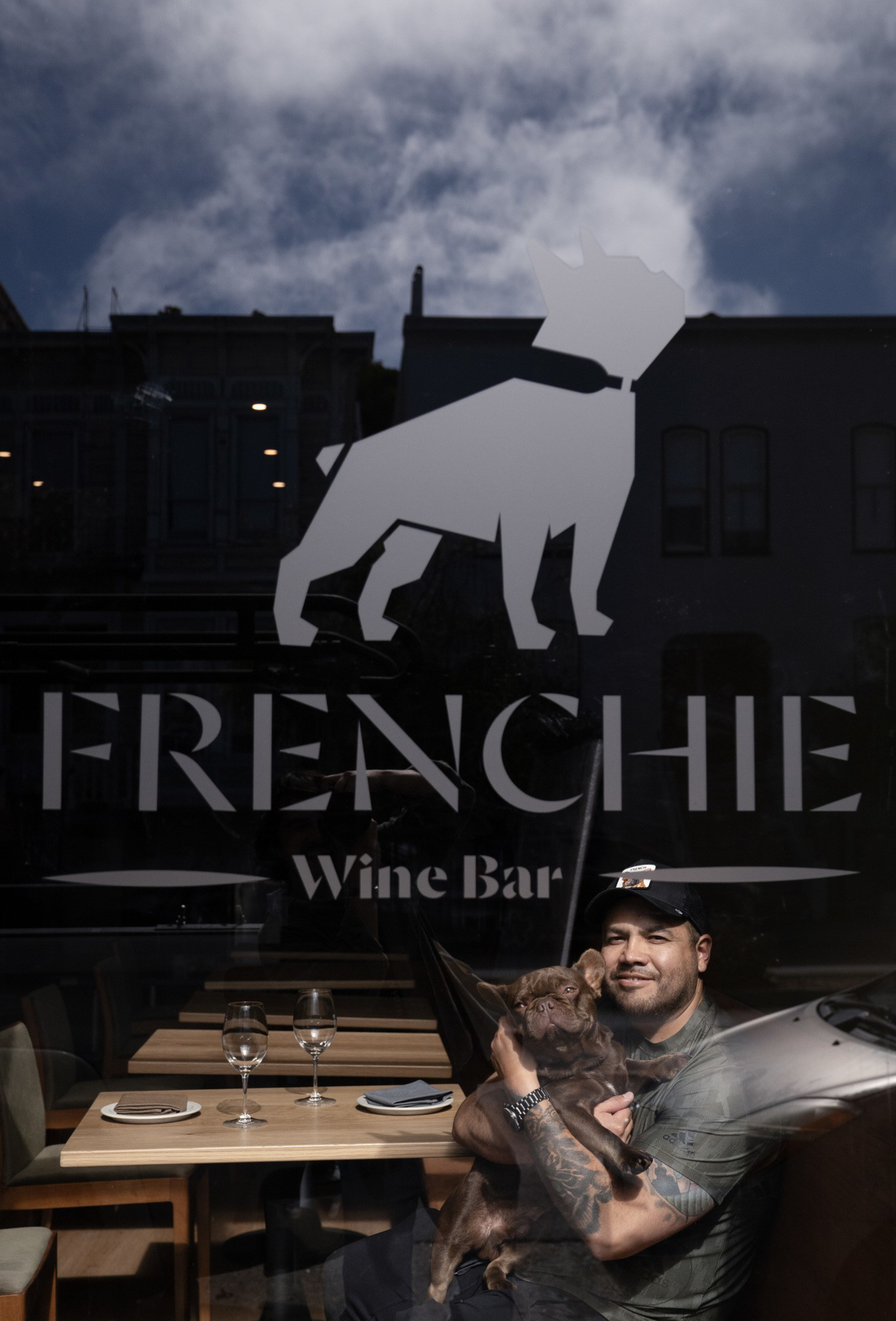 A man with tattoos and a cap sits inside a wine bar holding a small brown dog, seen through a window with a French Bulldog logo and "FRENCHIE Wine Bar" text.