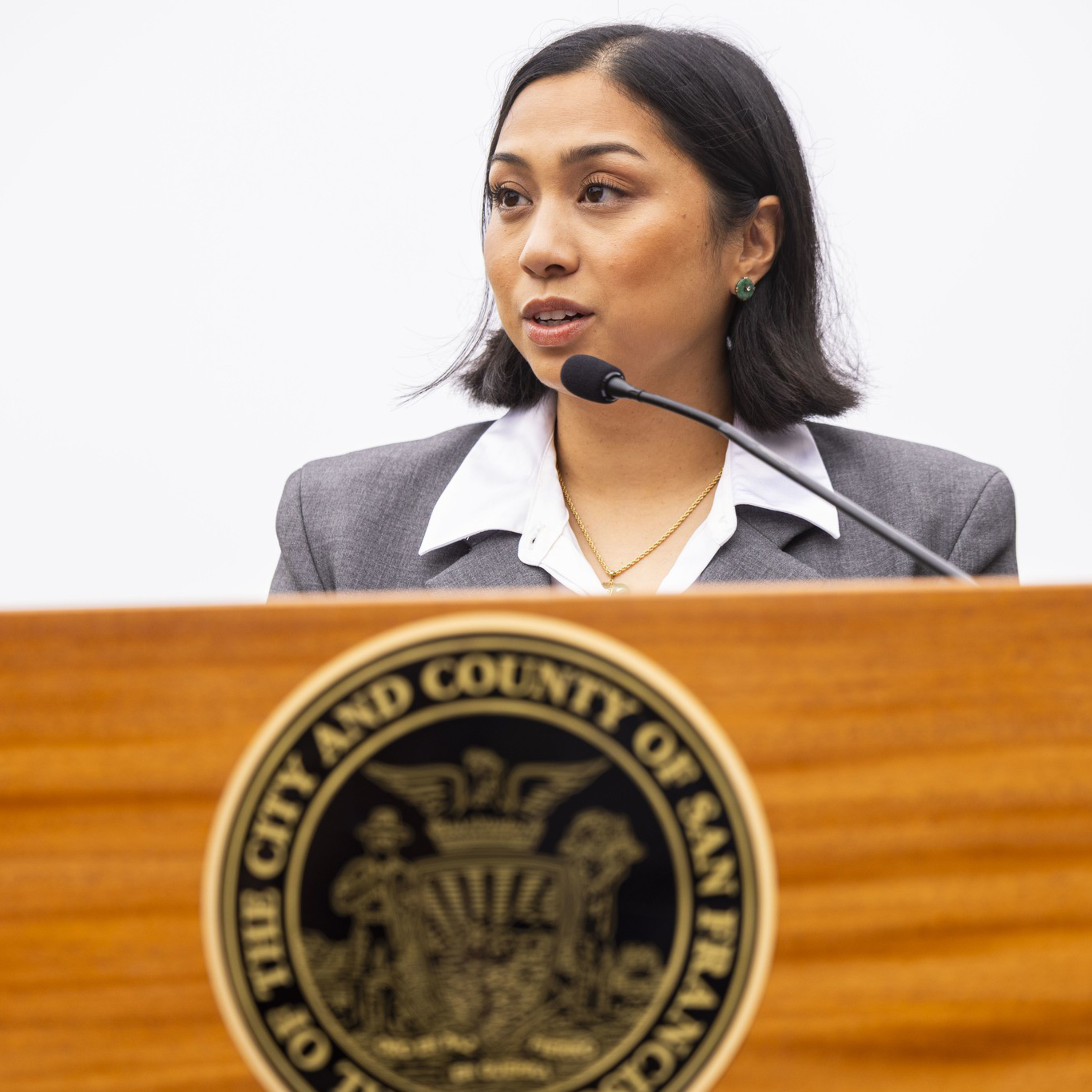 A woman in a gray blazer and white blouse speaks at a wooden podium with the seal of the City and County of San Francisco.