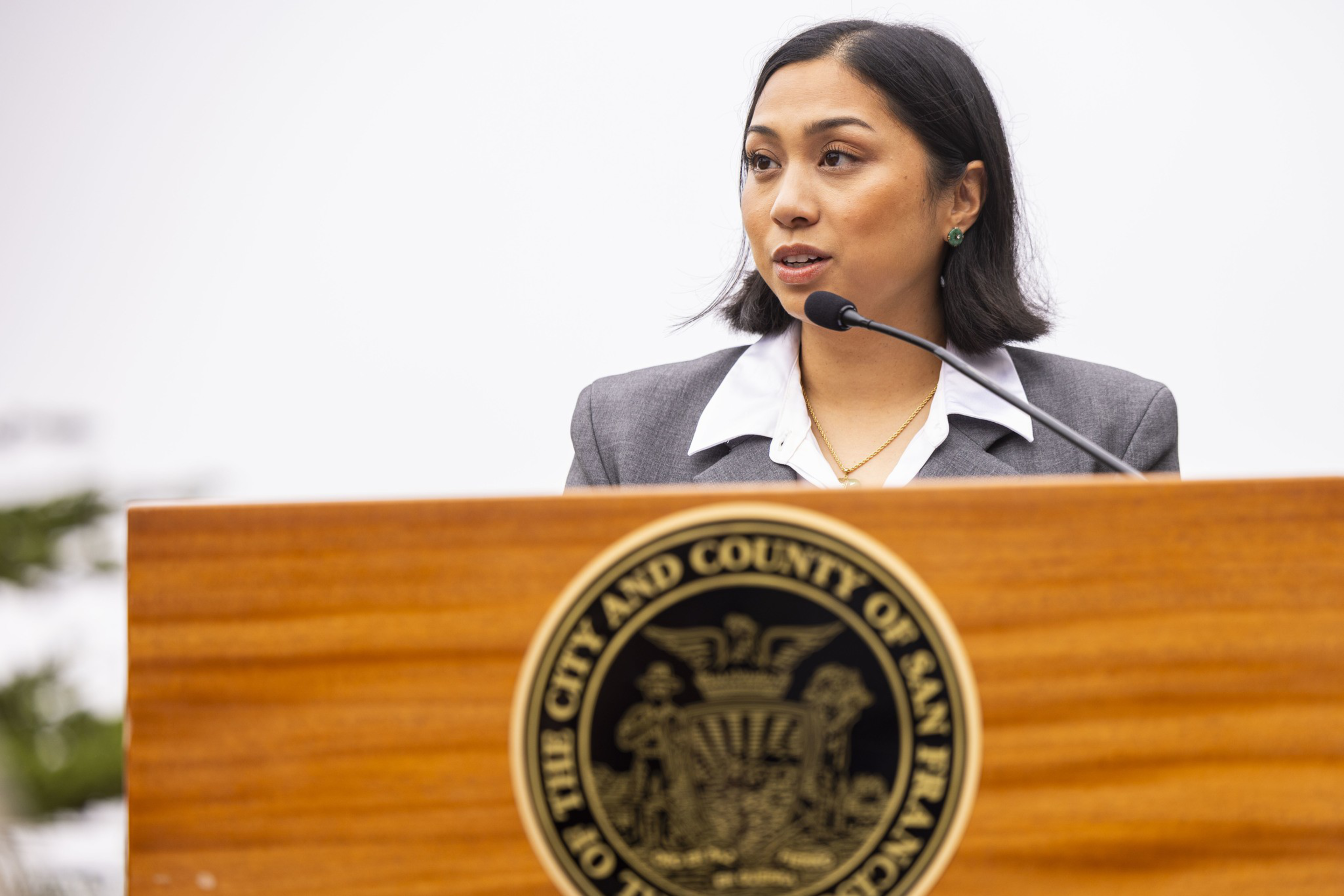 A woman in a gray blazer and white blouse speaks at a wooden podium with the seal of the City and County of San Francisco.