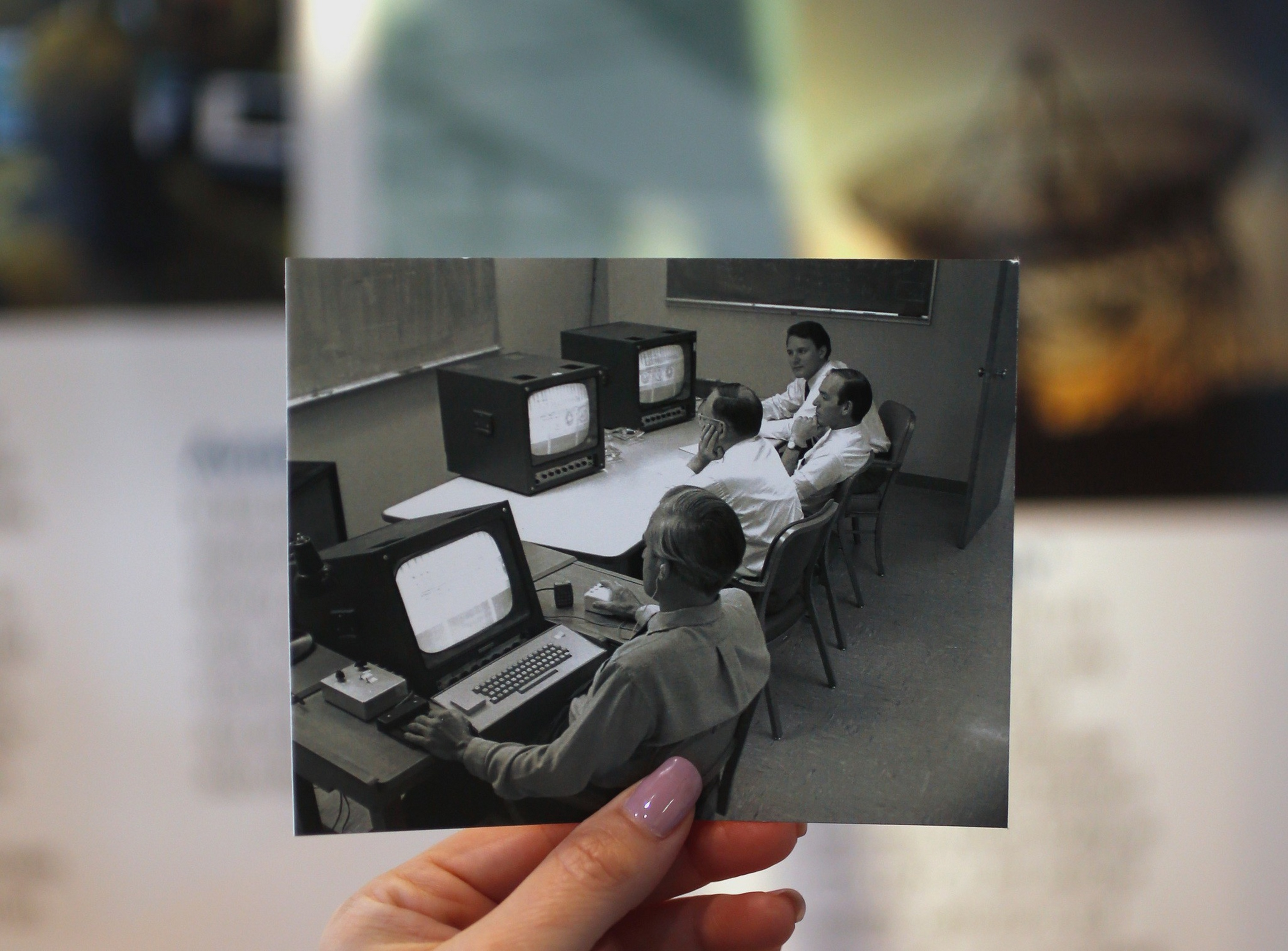 A hand with pink nail polish holds a black-and-white photo of four men in a room, using old computers and terminals from the mid-20th century.