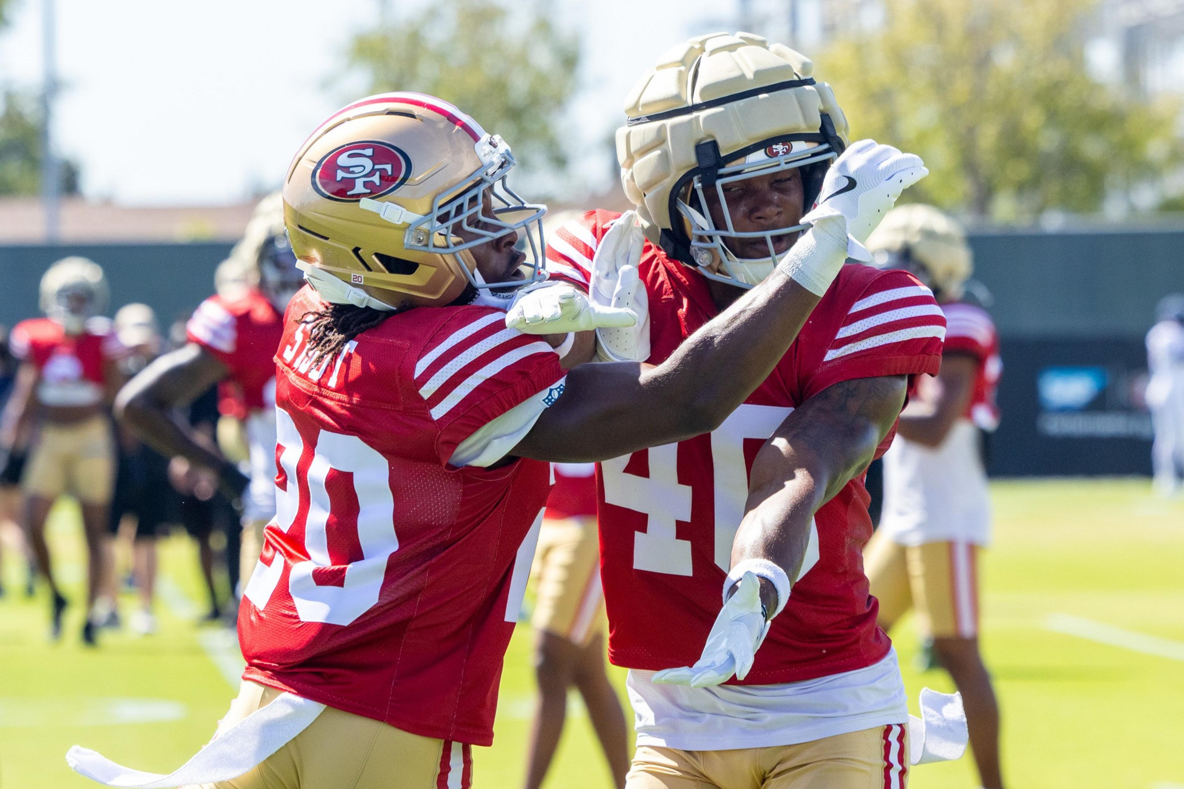 Two football players in red and gold San Francisco 49ers practice uniforms are interacting, with one player’s hand near the other’s face.