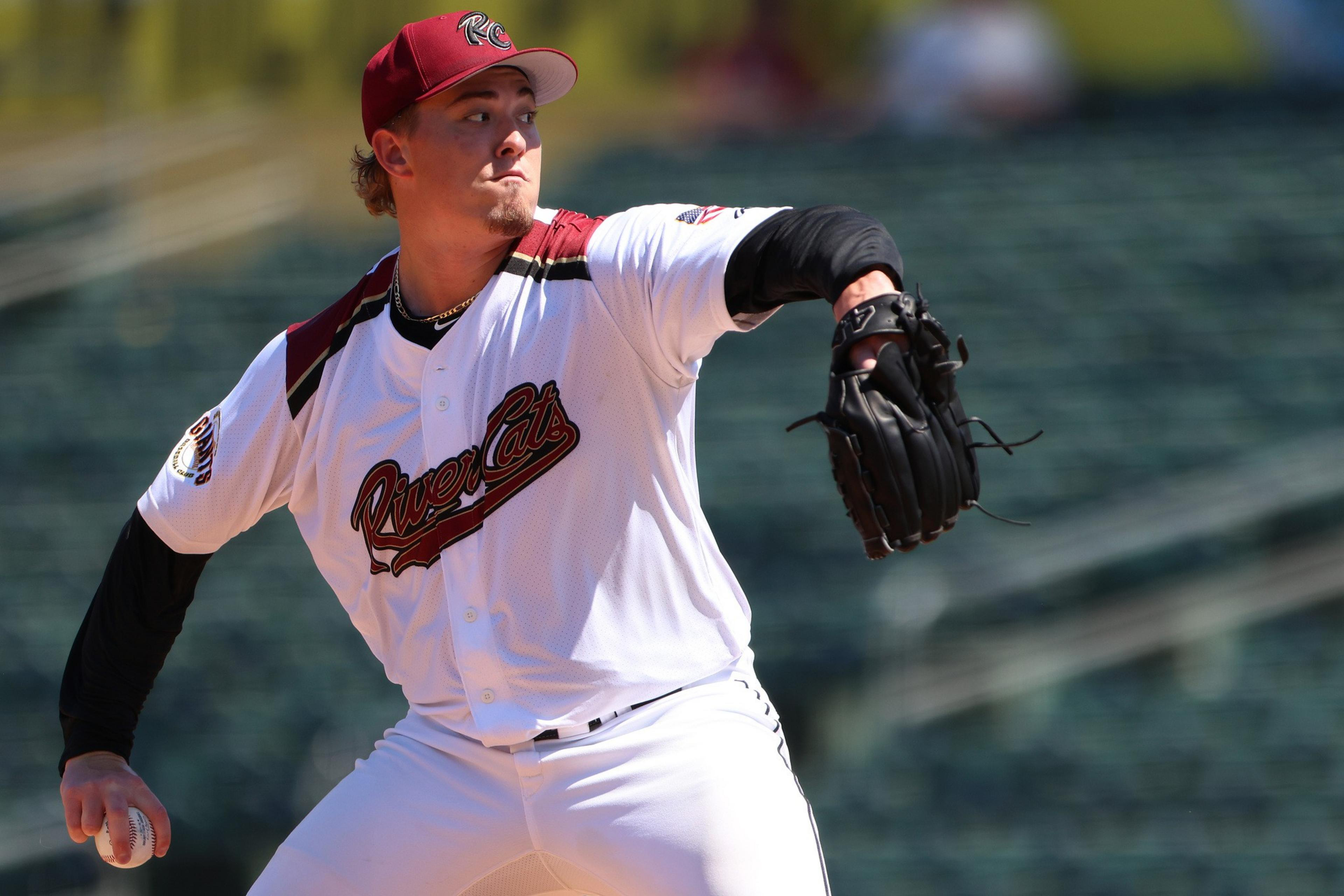 A baseball pitcher in a white River Cats uniform and red cap is winding up to throw a pitch in an empty stadium.