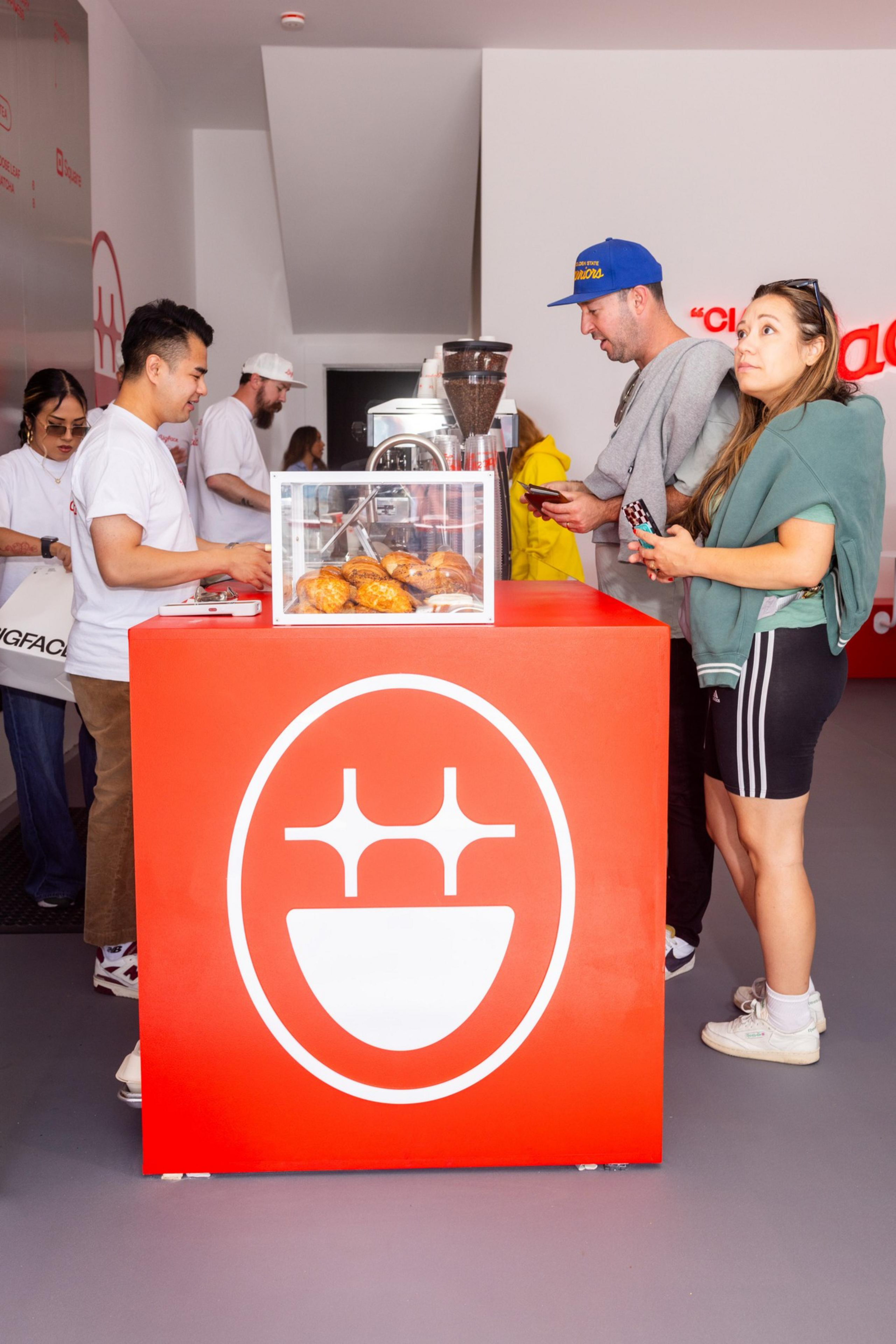 People stand at a red counter in a cafe, featuring pastries in a glass case and a coffee grinder. The logo on the counter resembles a smiling face.