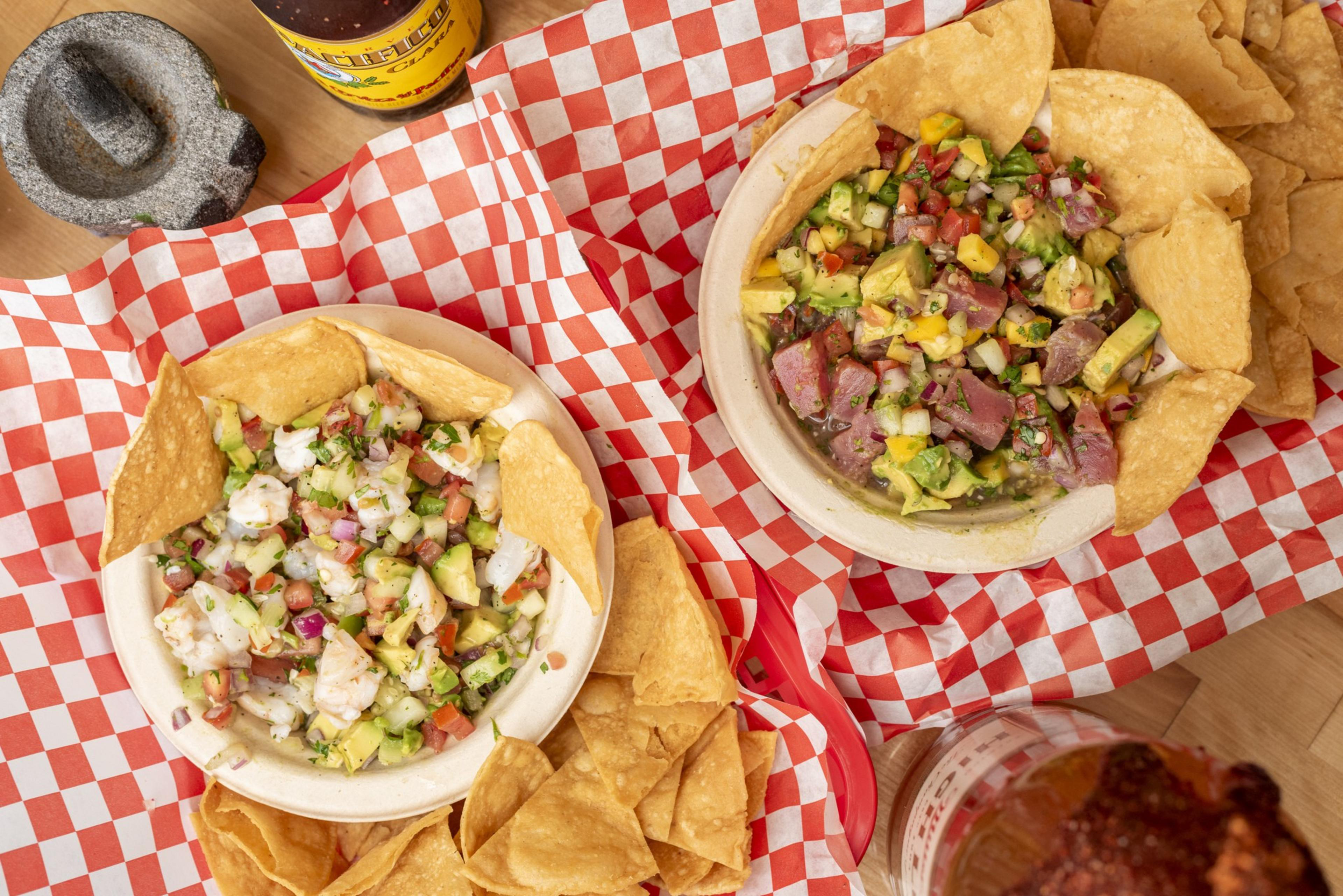 The image shows two bowls of vibrant ceviche with diced seafood and vegetables, surrounded by crispy tortilla chips, set on red and white checkered paper.