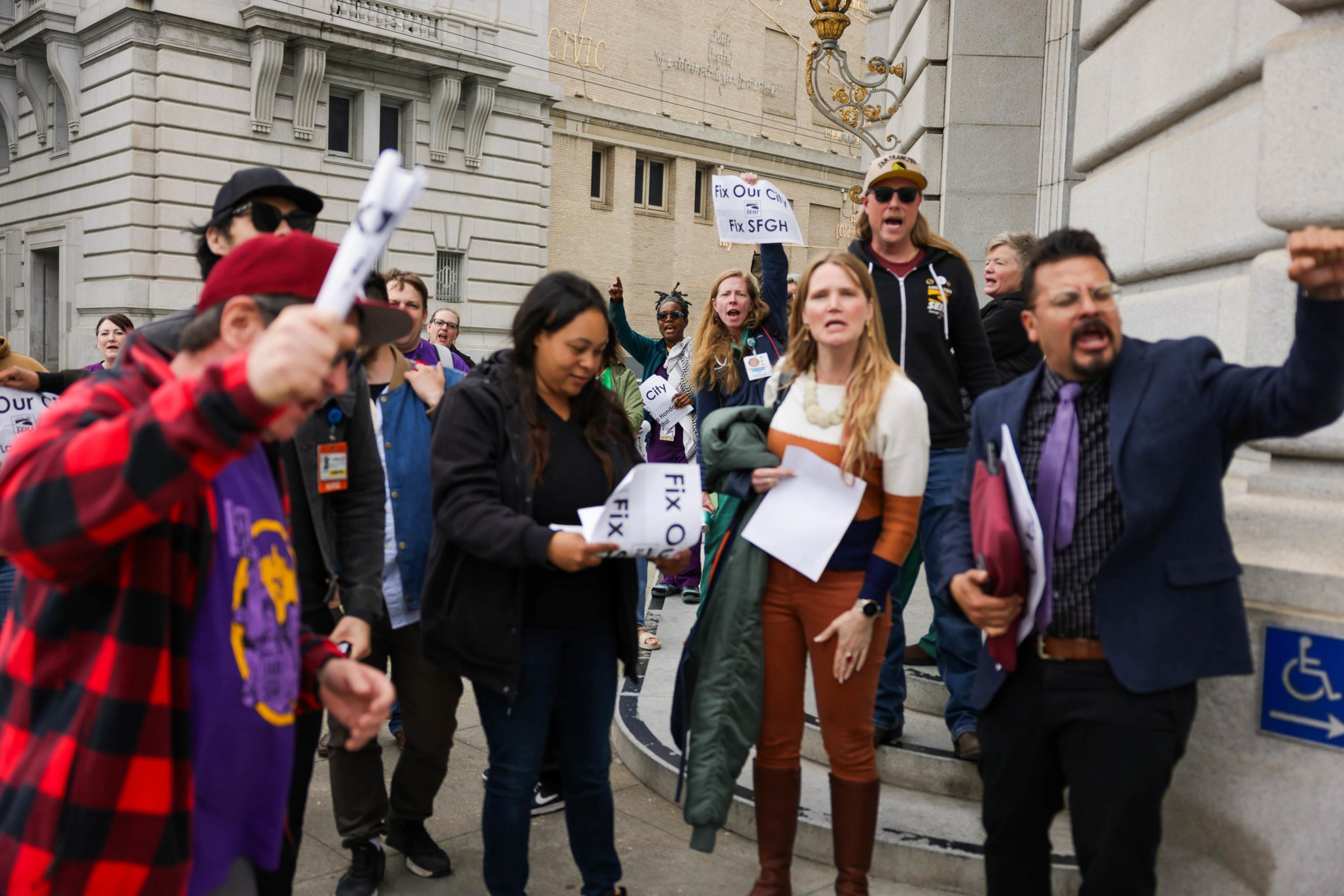 A group of people protest on steps, some holding signs and expressing vocal enthusiasm.