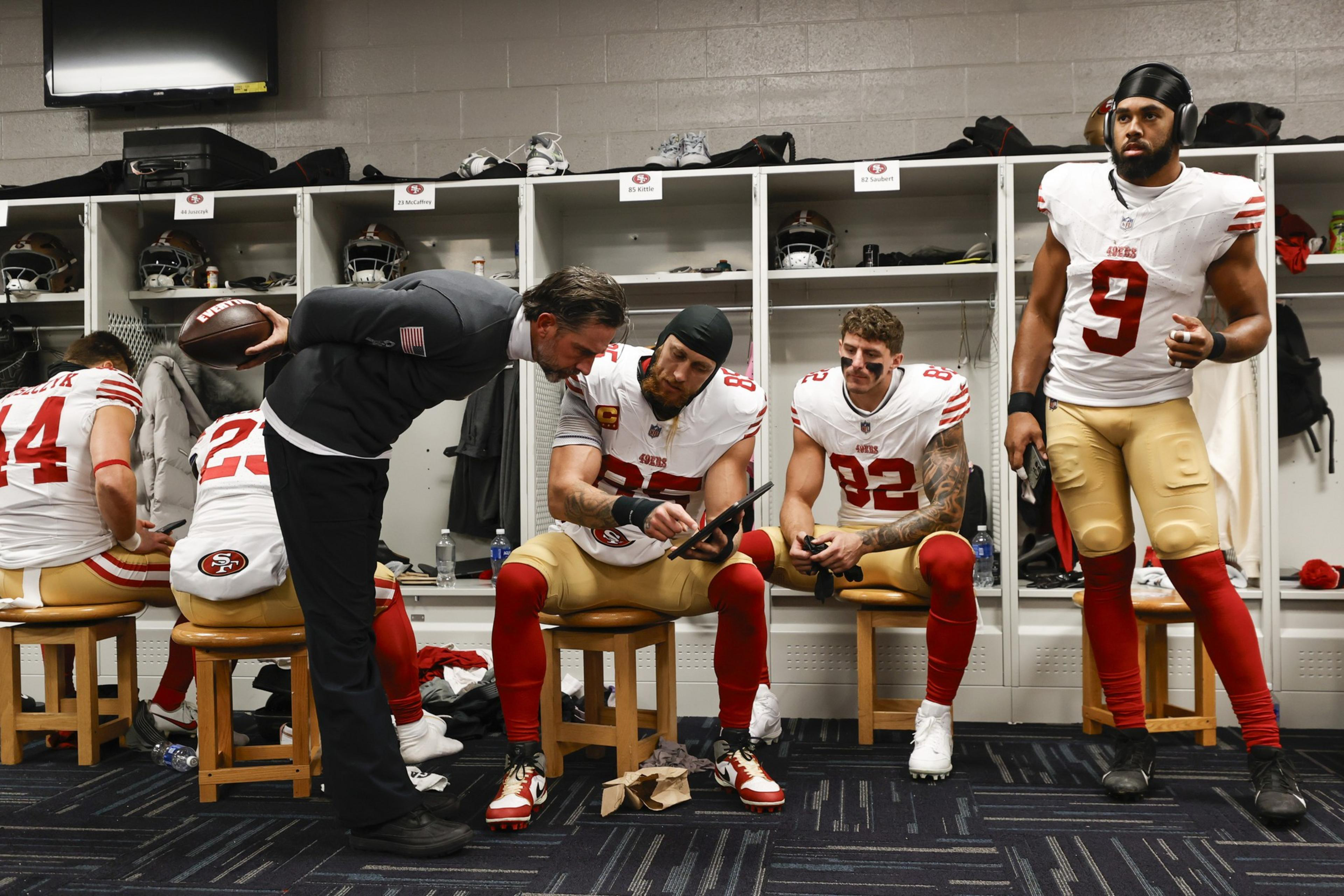Football players in a locker room wearing red and white jerseys are sitting on benches. They're looking at a tablet while another person, holding a football, leans over them.