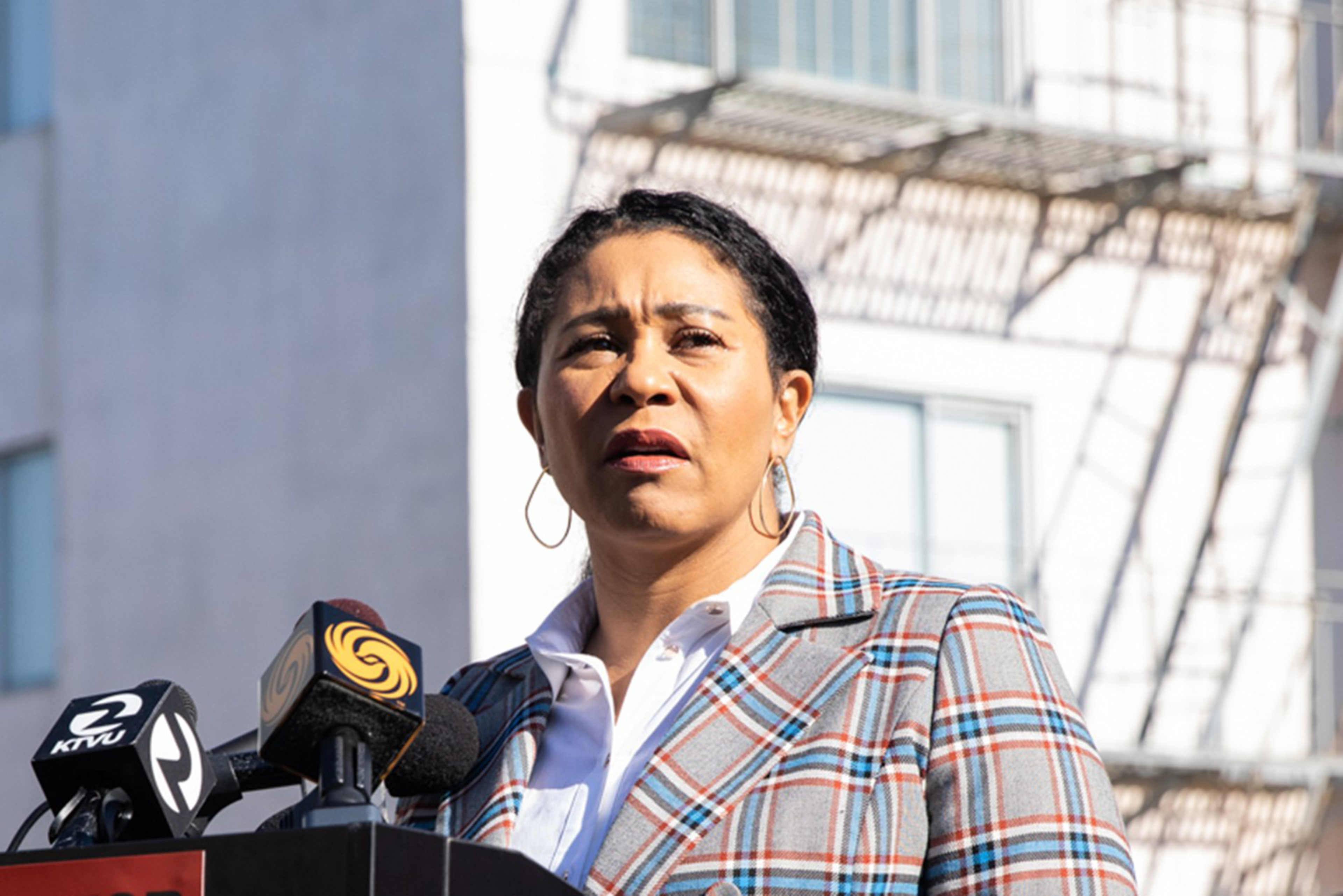 A woman wearing a plaid blazer speaks seriously at a podium with microphones, with a building and fire escape shadows in the background.