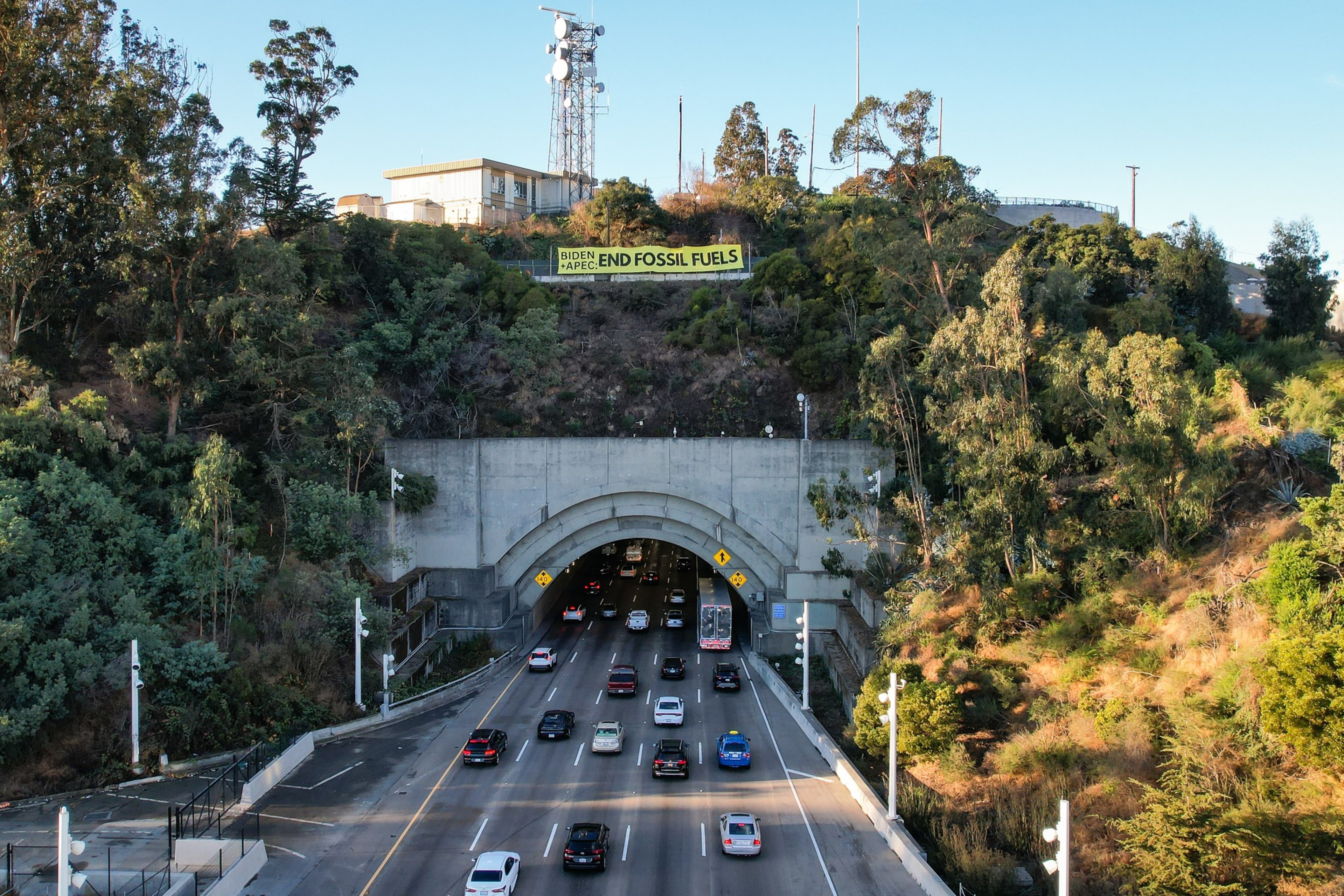 An aerial photo shows a large yellow banner reading "Biden and APEC: End Fossil Fuels" sitting above a tunnel with cars streaming through.