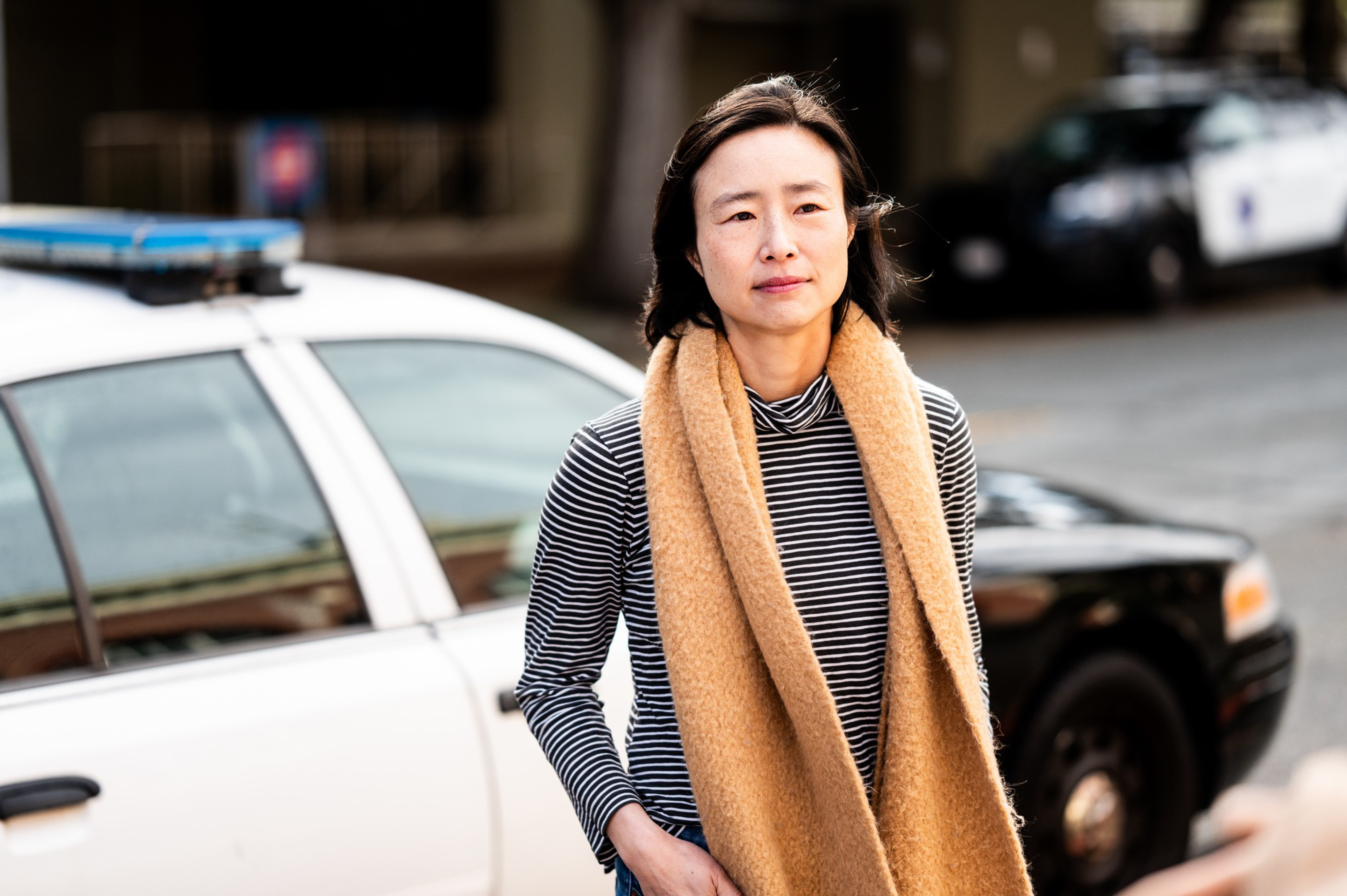 A woman stands in front of a police car, wearing a striped top and a tan scarf.