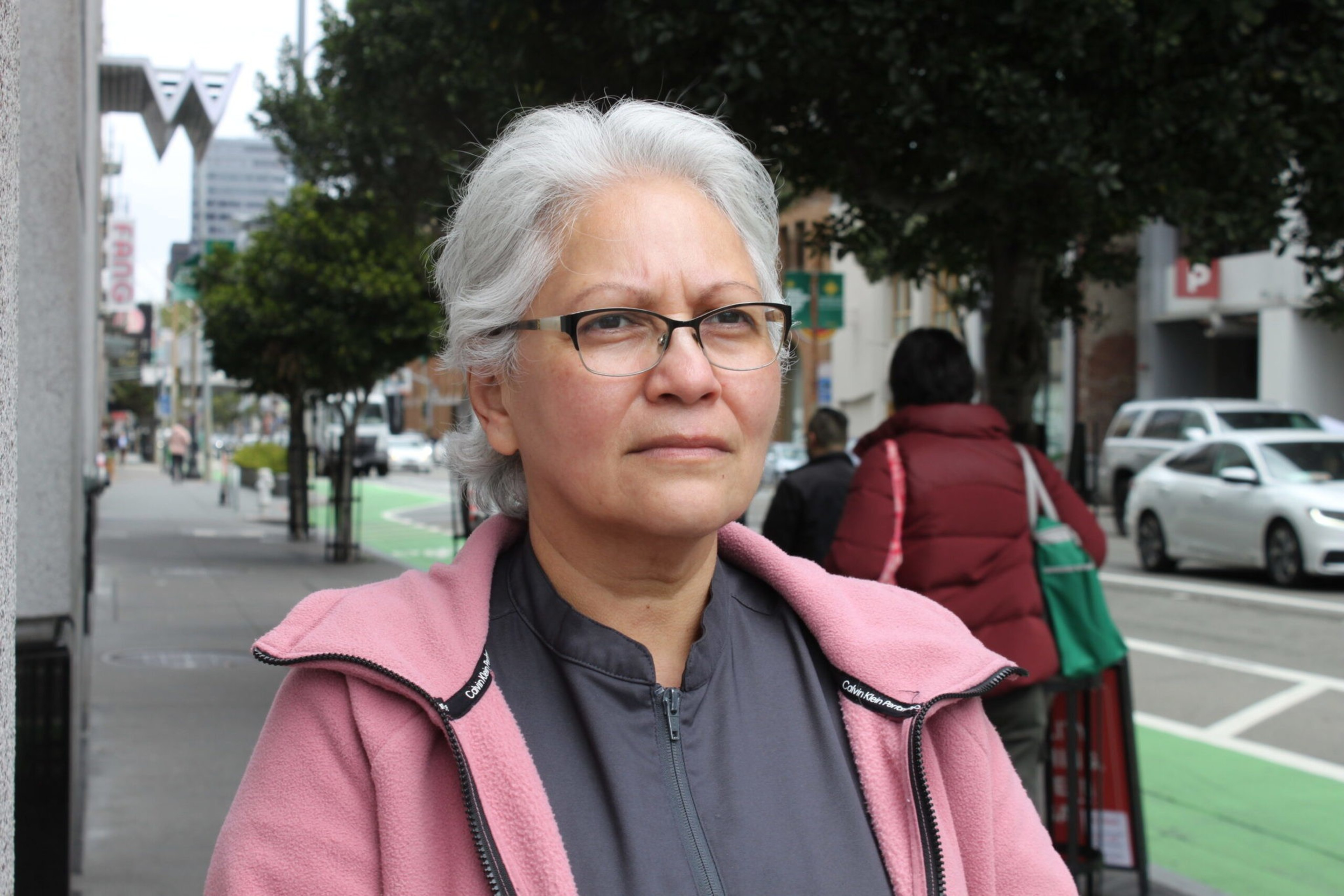 A woman with short white hair and glasses wears a pink jacket over a gray shirt, standing on a city sidewalk with trees and cars in the background.