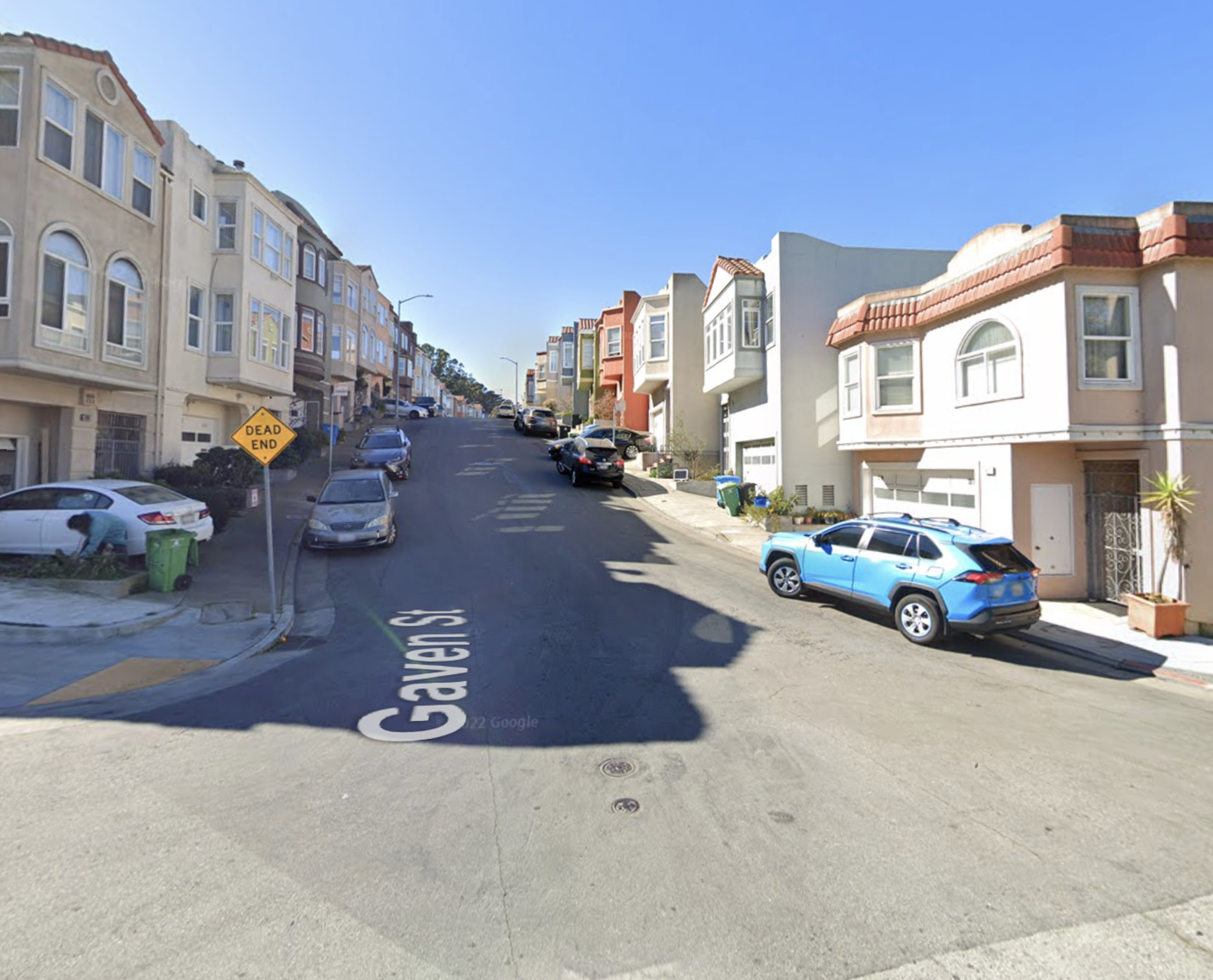 A steep residential street lined with parked cars and multi-story houses under a clear blue sky, featuring a "Dead End" sign on the left.