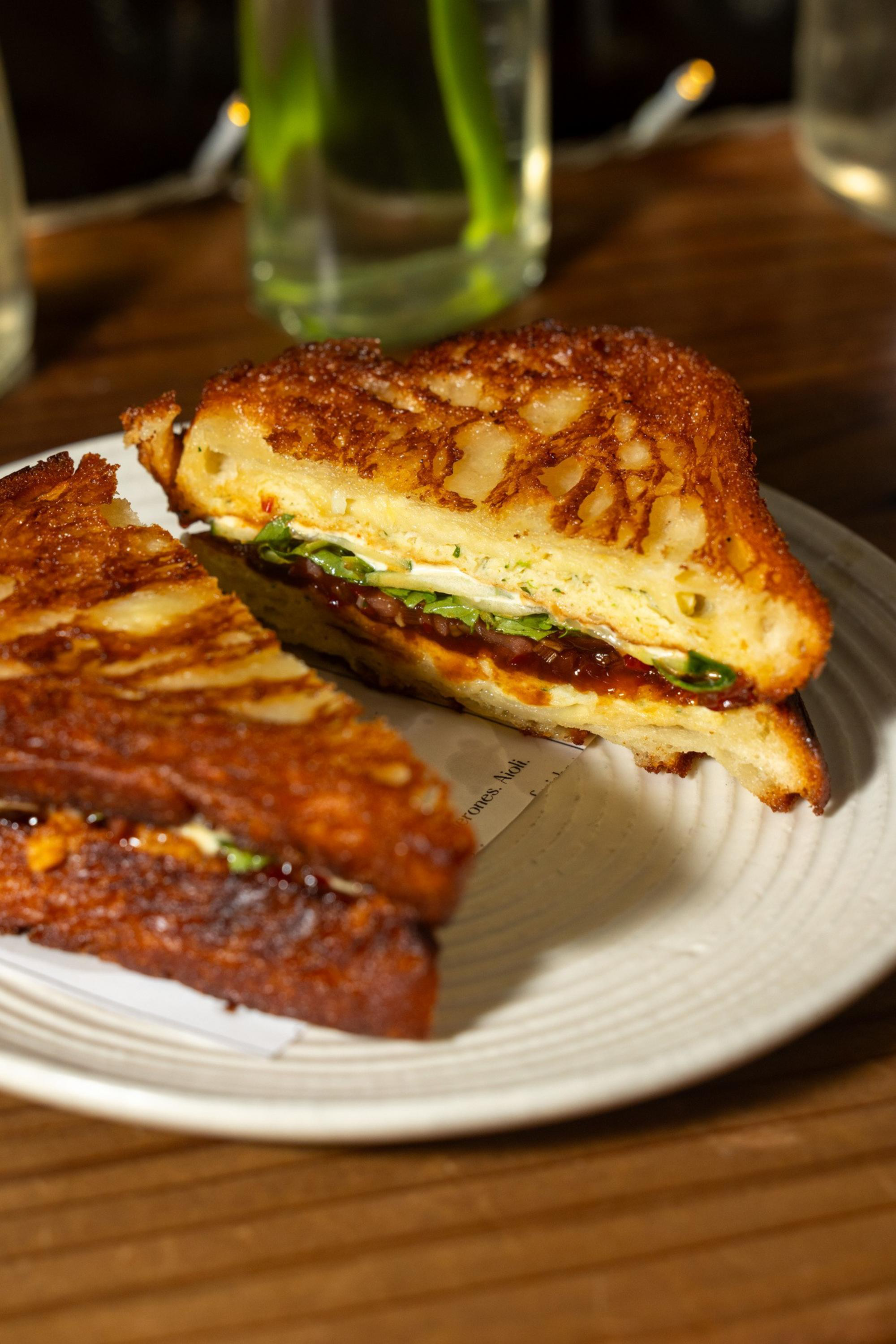 A toasted grilled cheese sandwich cut in half reveals melted cheese, greens, and a layer of red spread, served on a white plate on a wooden table.