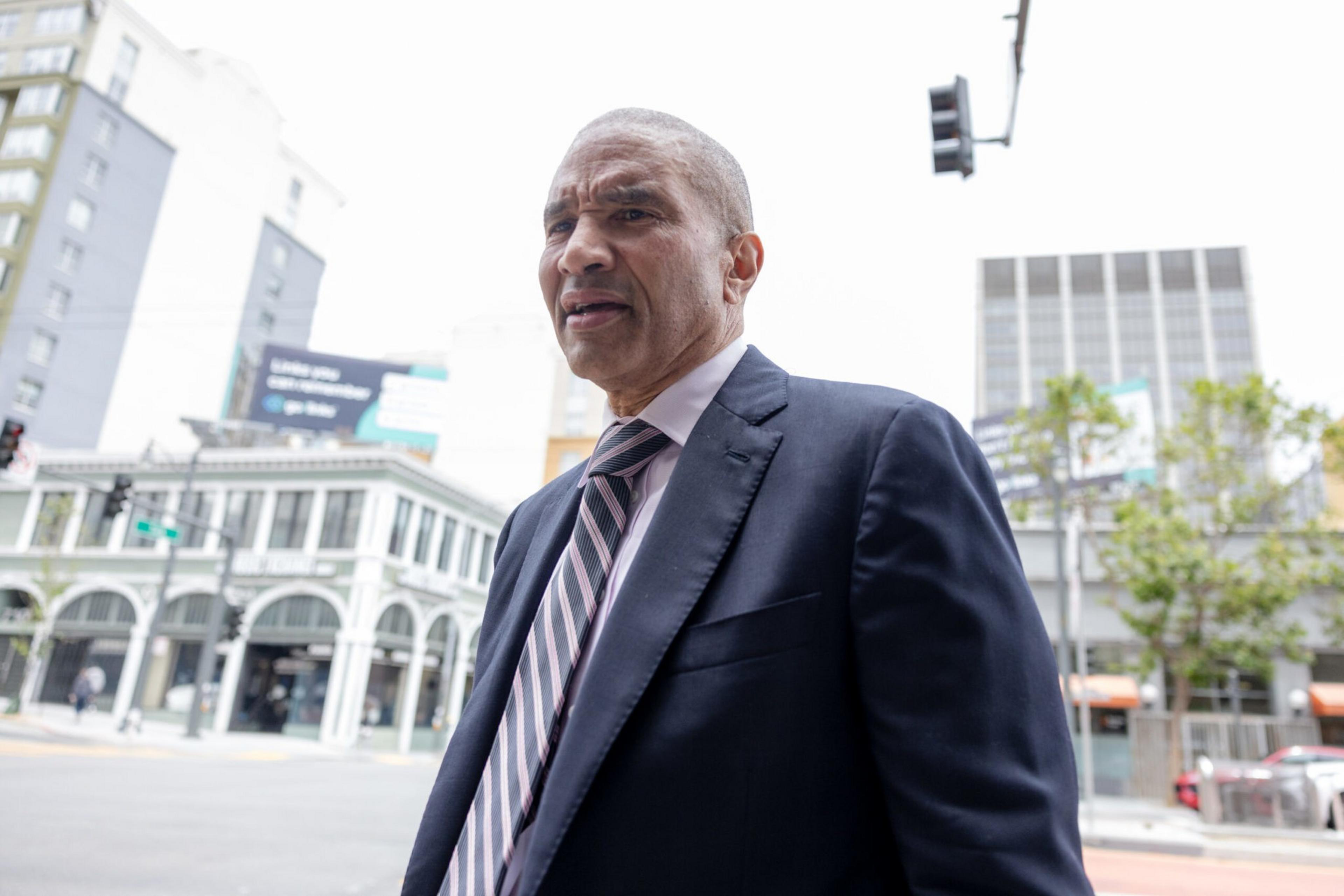 A man in a suit and striped tie stands on a city street with buildings and trees visible in the background under an overcast sky.