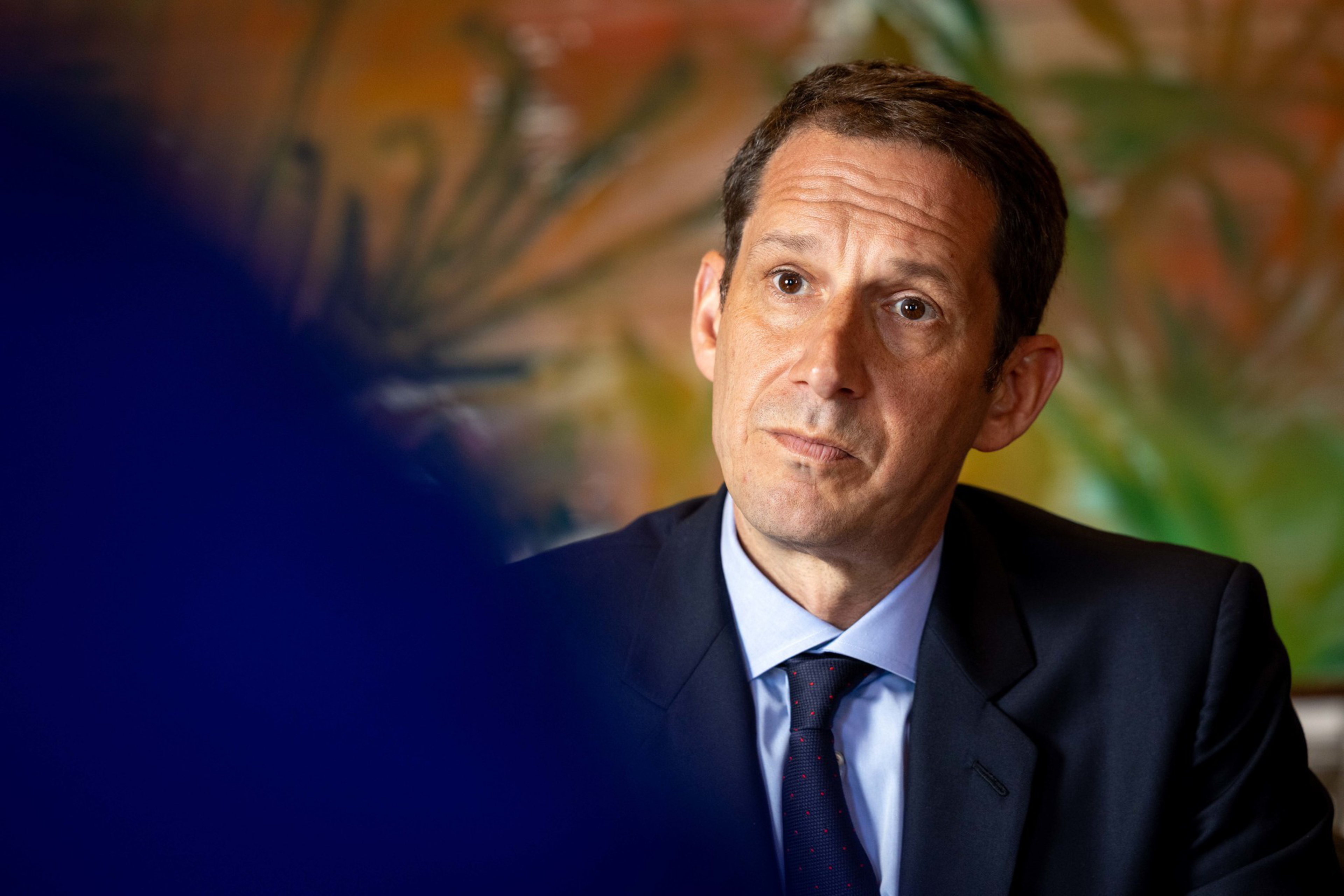 A man in a suit and tie looks thoughtfully to the side. The background is colorful and abstract, creating a contrast with his formal attire.