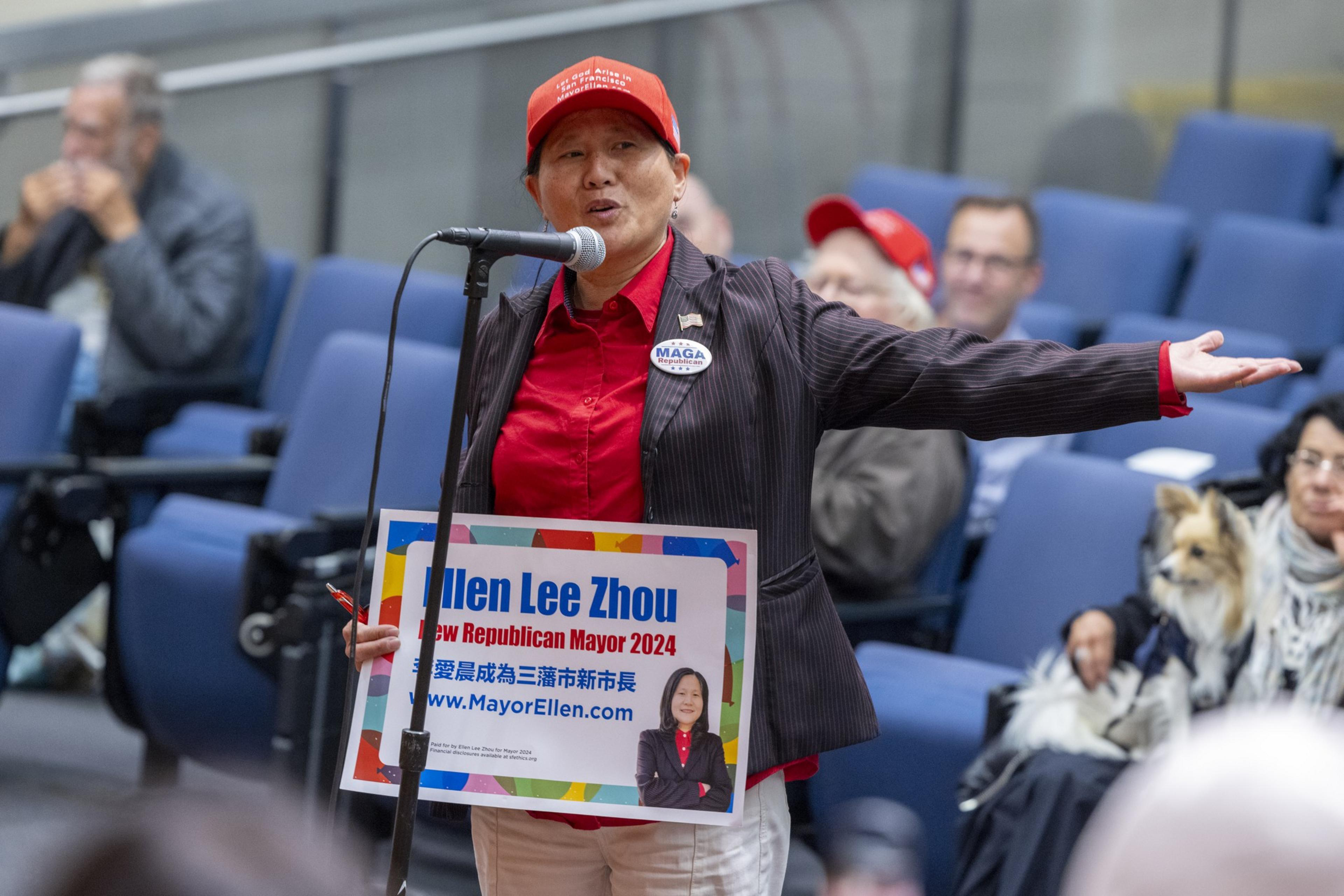 A woman in a red cap and shirt speaks into a microphone, holding a campaign sign for Ellen Lee Zhou, in a room with blue seats and an audience.