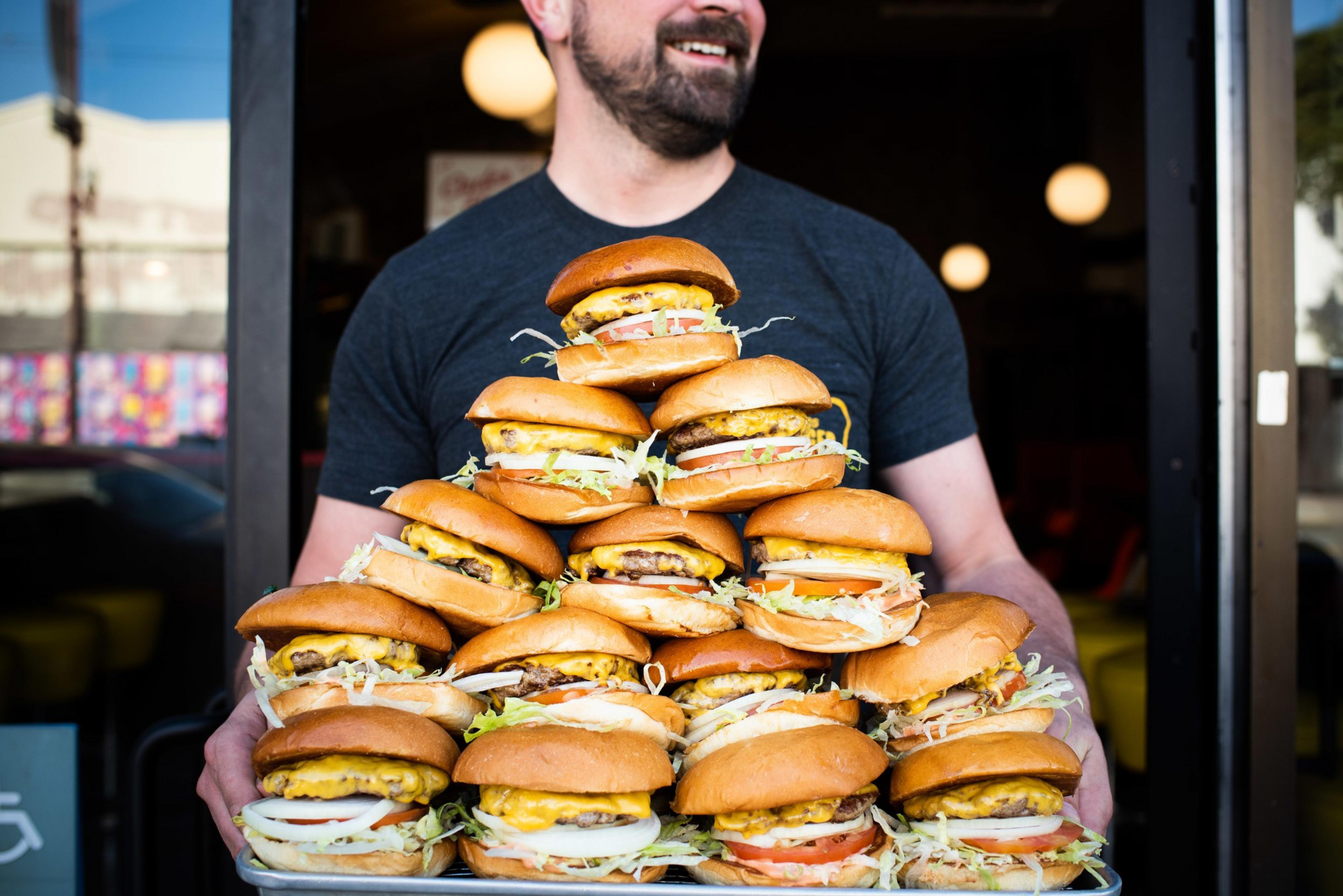 A man stands smiling, holding a large tray with a pyramid of twenty cheeseburgers, each stacked with lettuce, tomato, and cheese, outside a restaurant entrance.