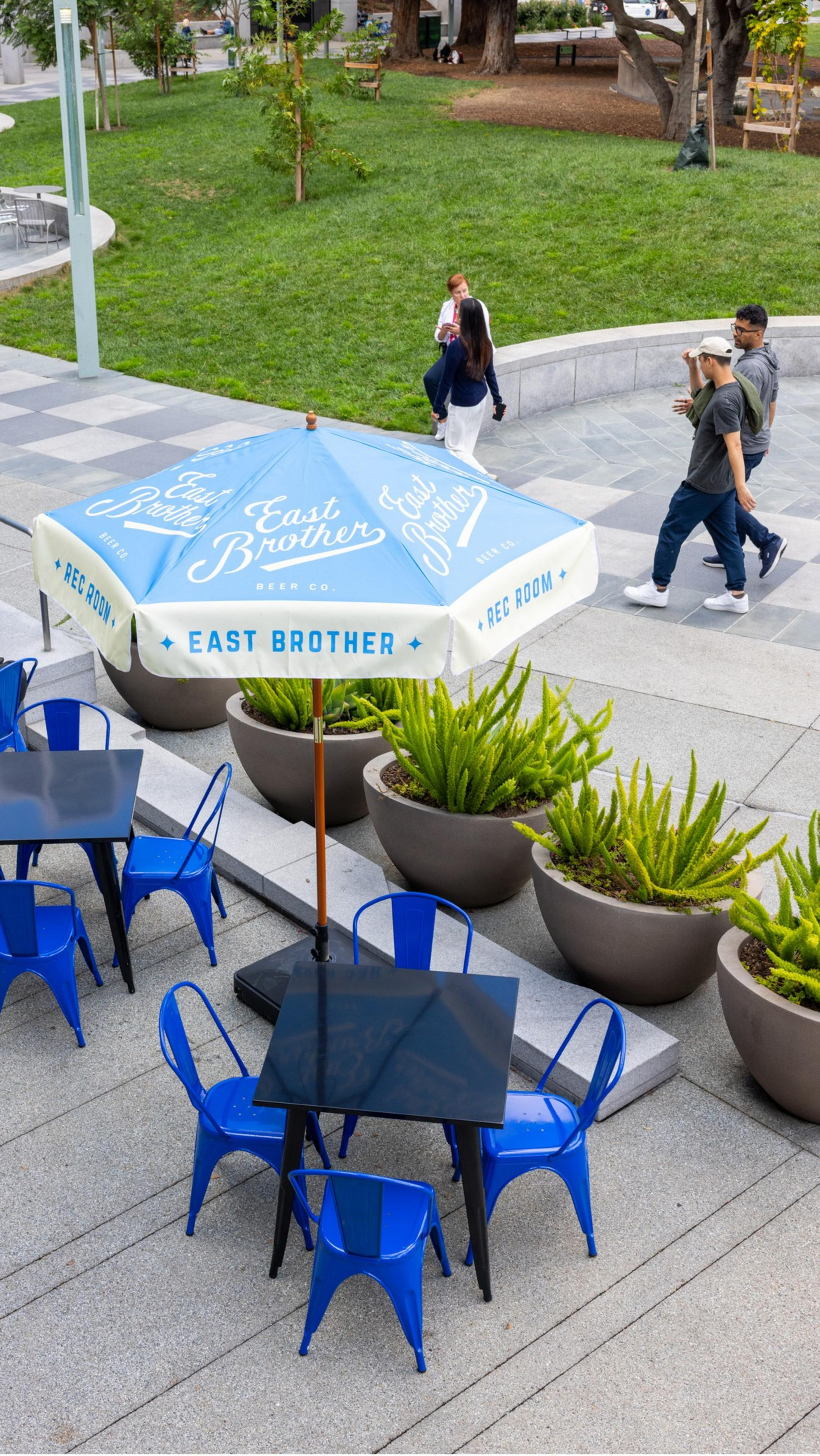 Blue chairs and tables surround a patio umbrella with "East Brother" branding beside large potted plants, near a grassy area with walking pedestrians.