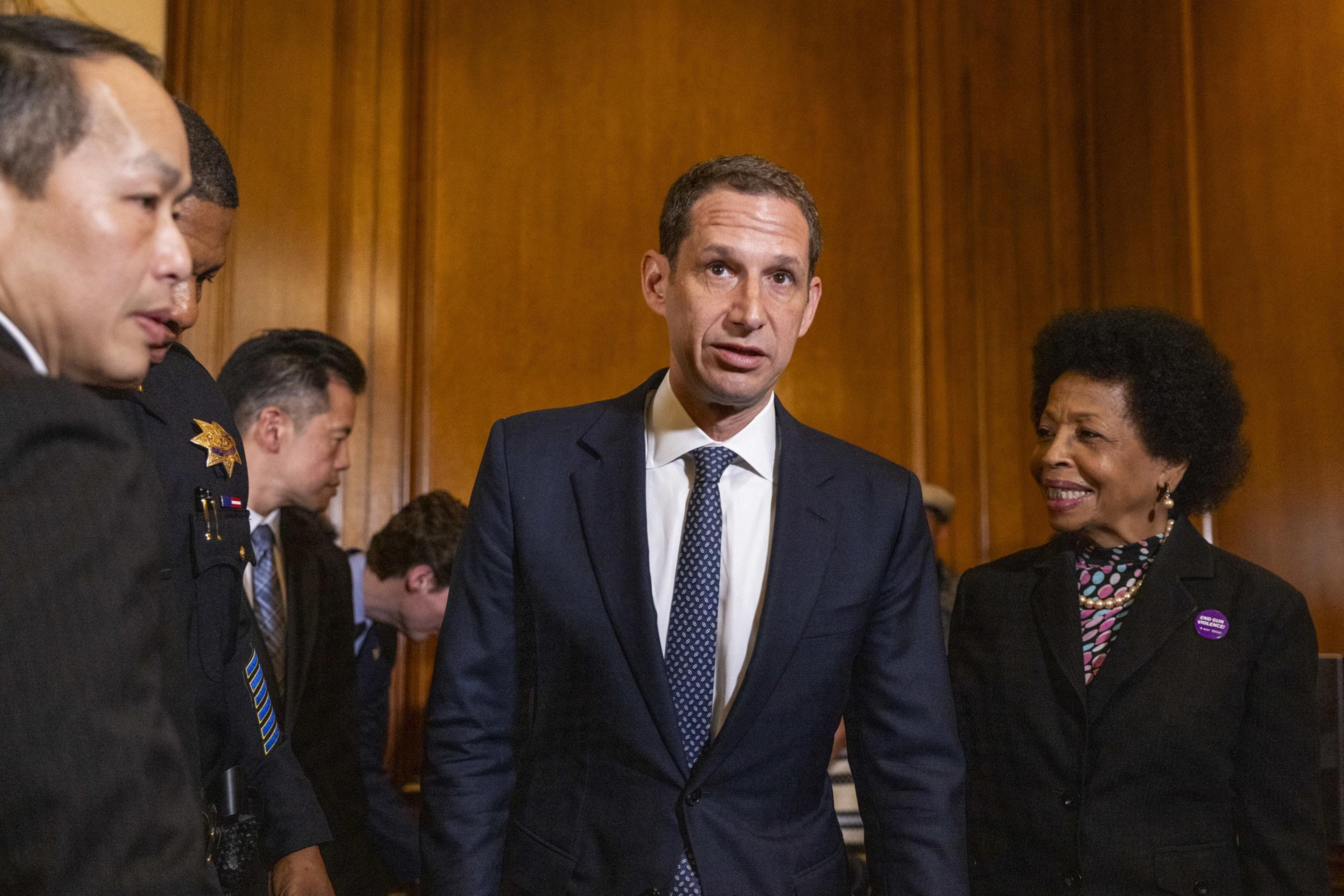 A group of people in formal attire gather in a wood-paneled room. A man in a suit stands in the center, while others interact around him, smiling.