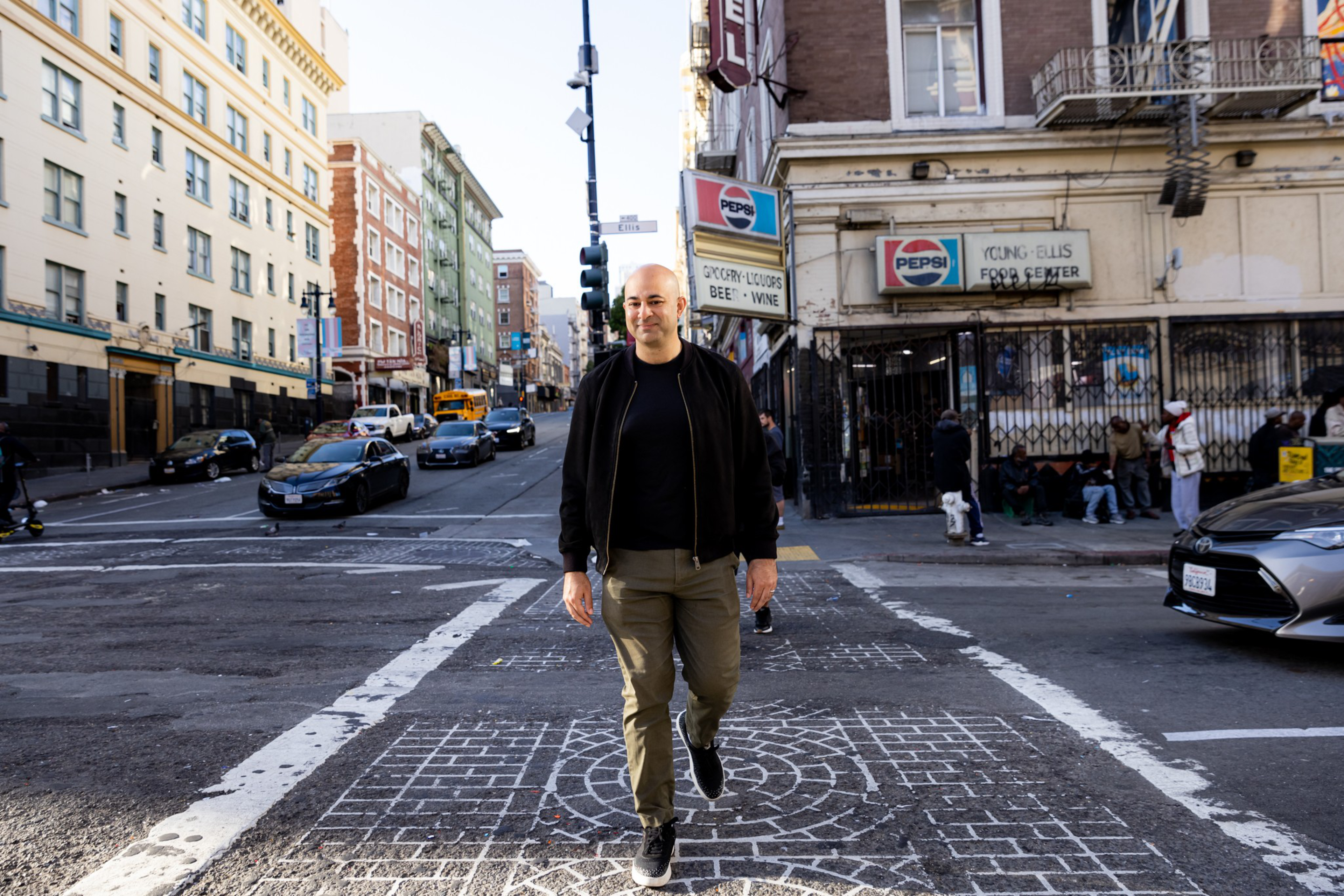 A man wearing a black jacket and olive pants walks across a city street with buildings, cars, and people near a store labeled “Young-Ellis Food Center.”